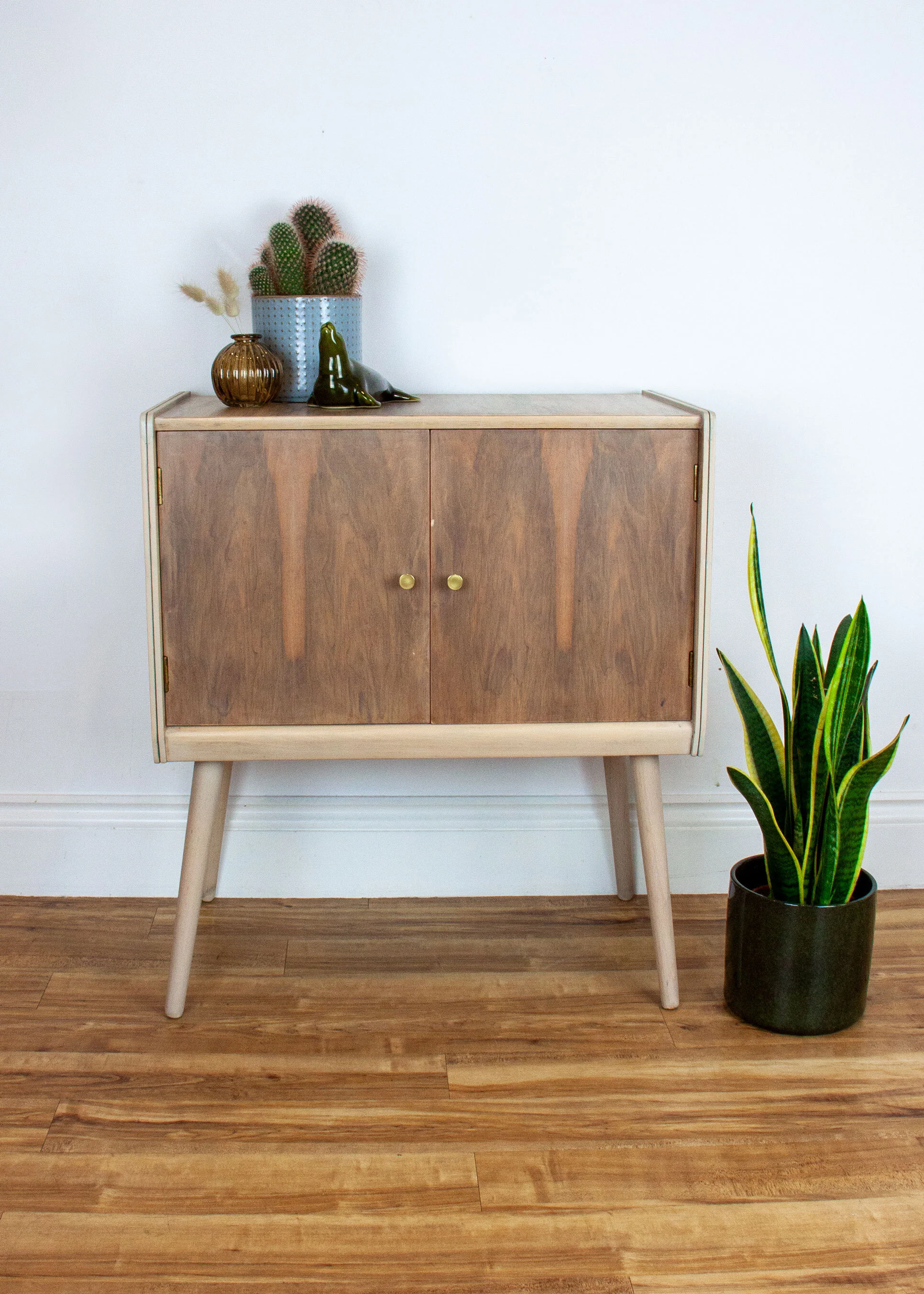 A mid-century modern wooden cabinet with angled legs, placed against a plain white wall. On top of the cabinet, there are decorative items including a blue textured planter with a cactus, a small brown vase, and a dark green ceramic animal figure. To