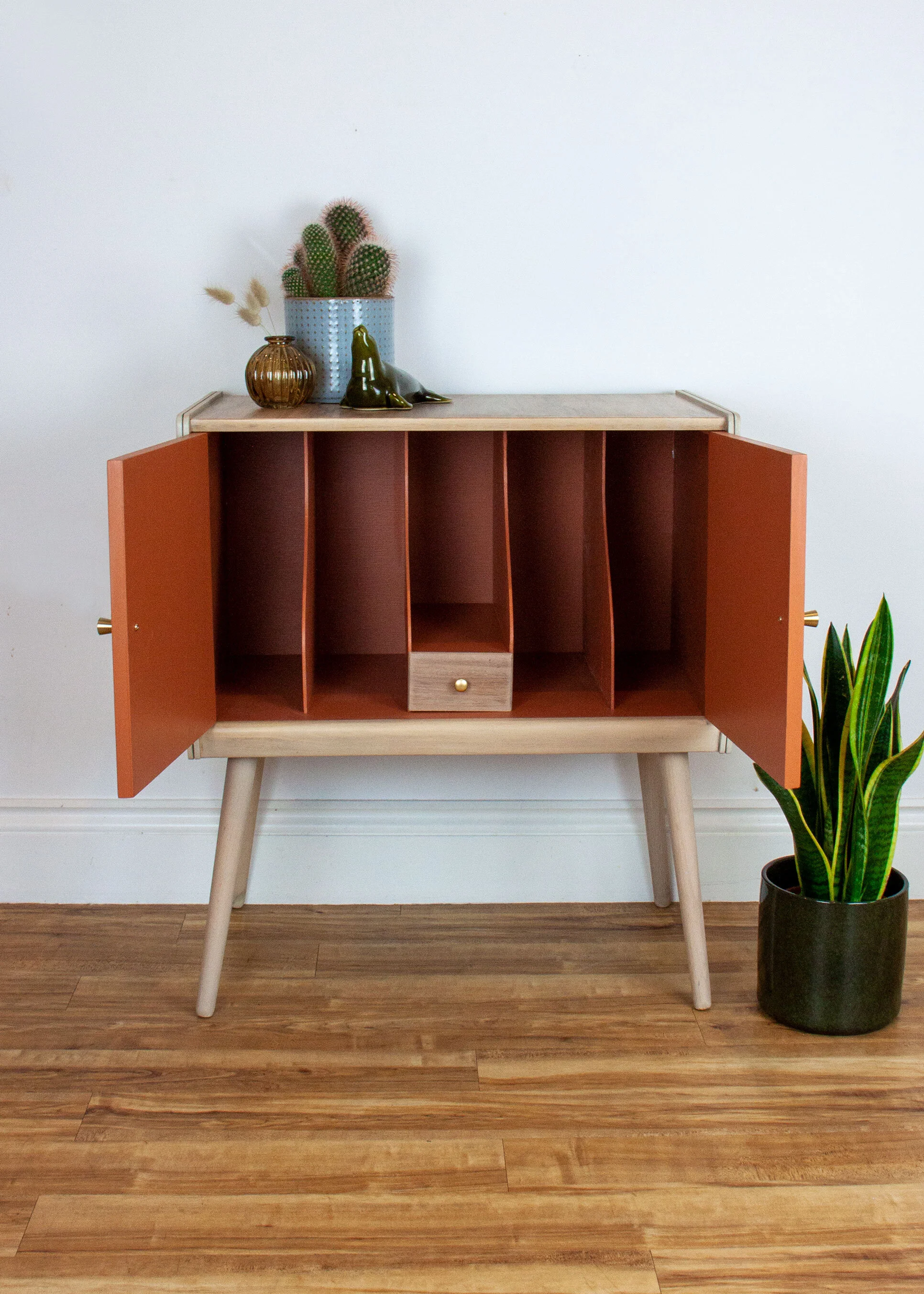A mid-century modern cabinet with open orange doors, set on four angled legs, on a wooden floor against a white wall. The top of the cabinet holds potted cactus, decorative vases, and figurines. A potted snake plant is on the right side on the floor.