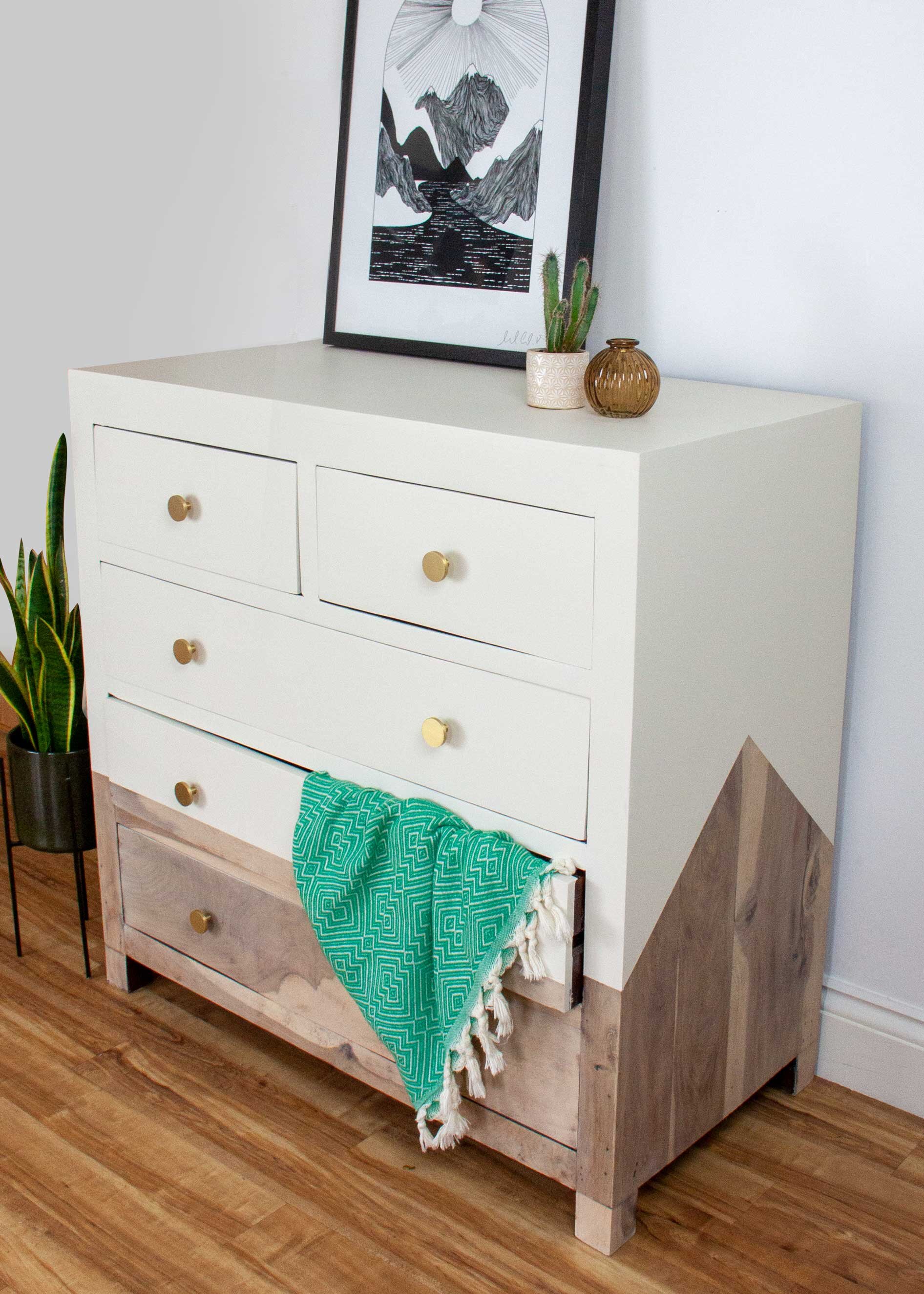 A white and natural wood dresser with gold knobs, topped with a framed black and white mountain landscape art, small potted cactus, and decorative vases. A teal patterned throw hangs on the bottom drawer. A tall green plant is beside the dresser, and