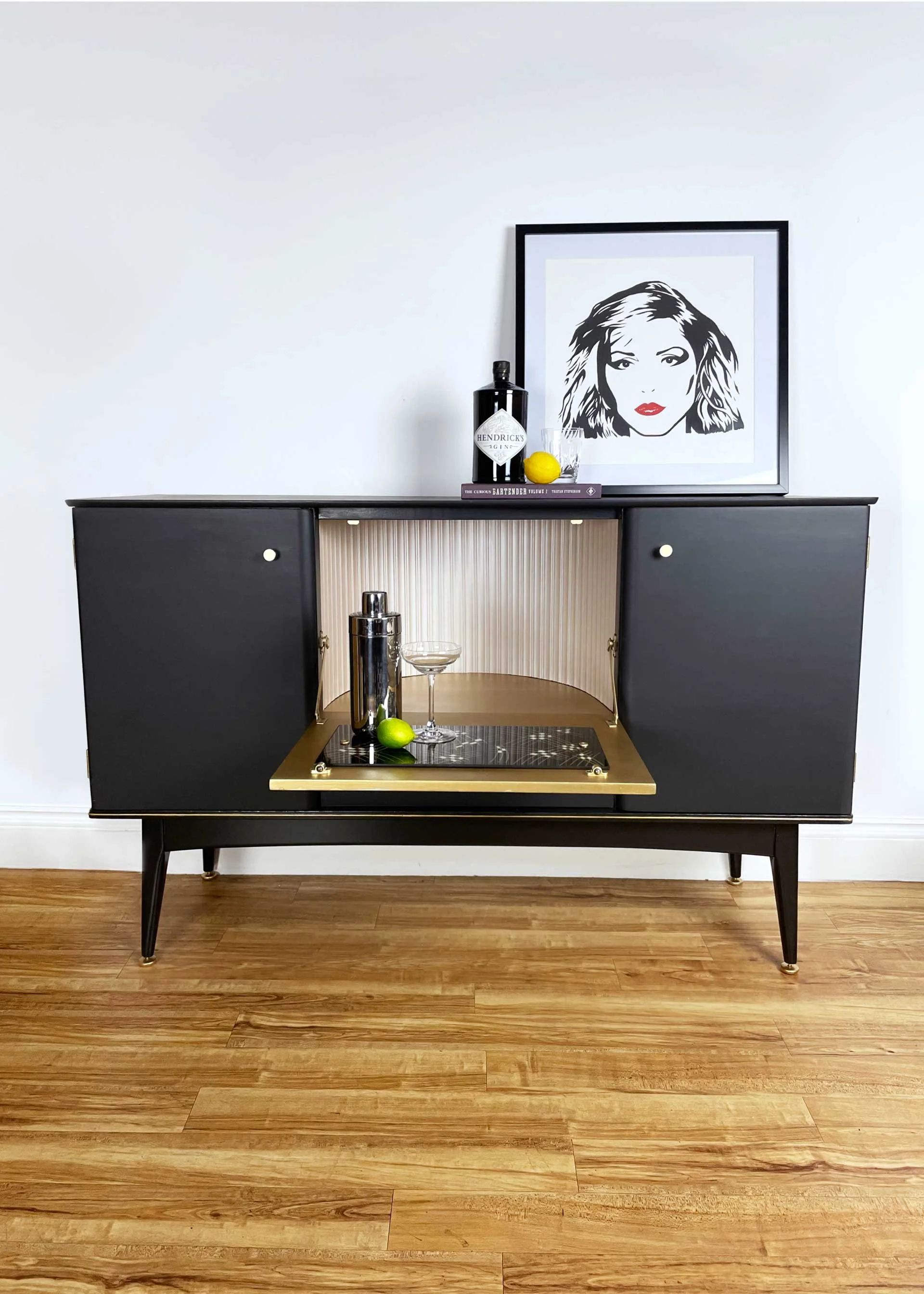 A black vintage-style bar cabinet with open middle section, decorated with a framed black-and-white portrait of a woman with red lipstick, a bottle of Hendrick's gin, a lemon, a glass, a silver cocktail shaker, and a green lime; some books and a glas