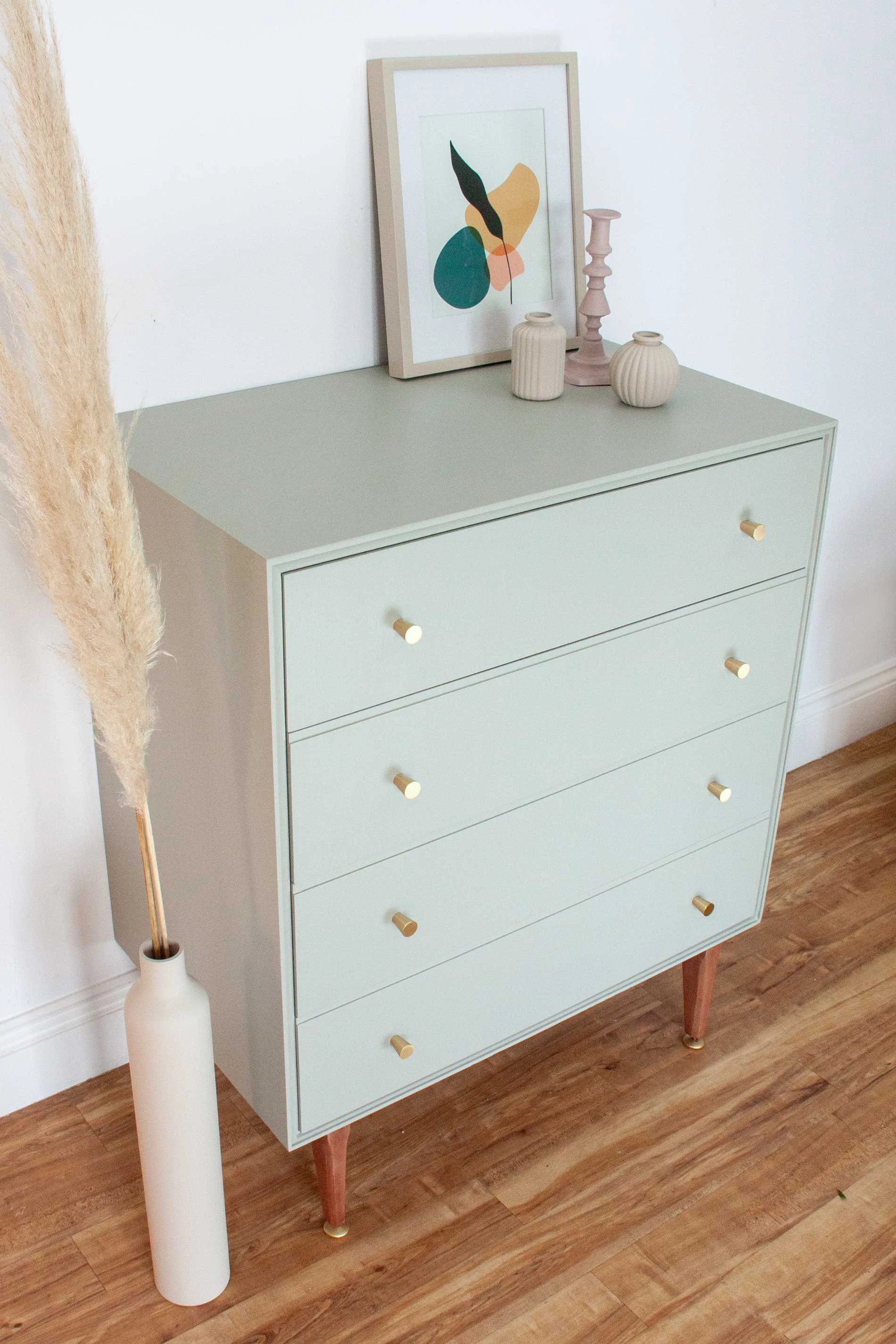 A light green dresser with five drawers and wooden tapered legs, decorated with a framed abstract art print, three vases, and a pink candlestick, set on wooden flooring against a white wall.