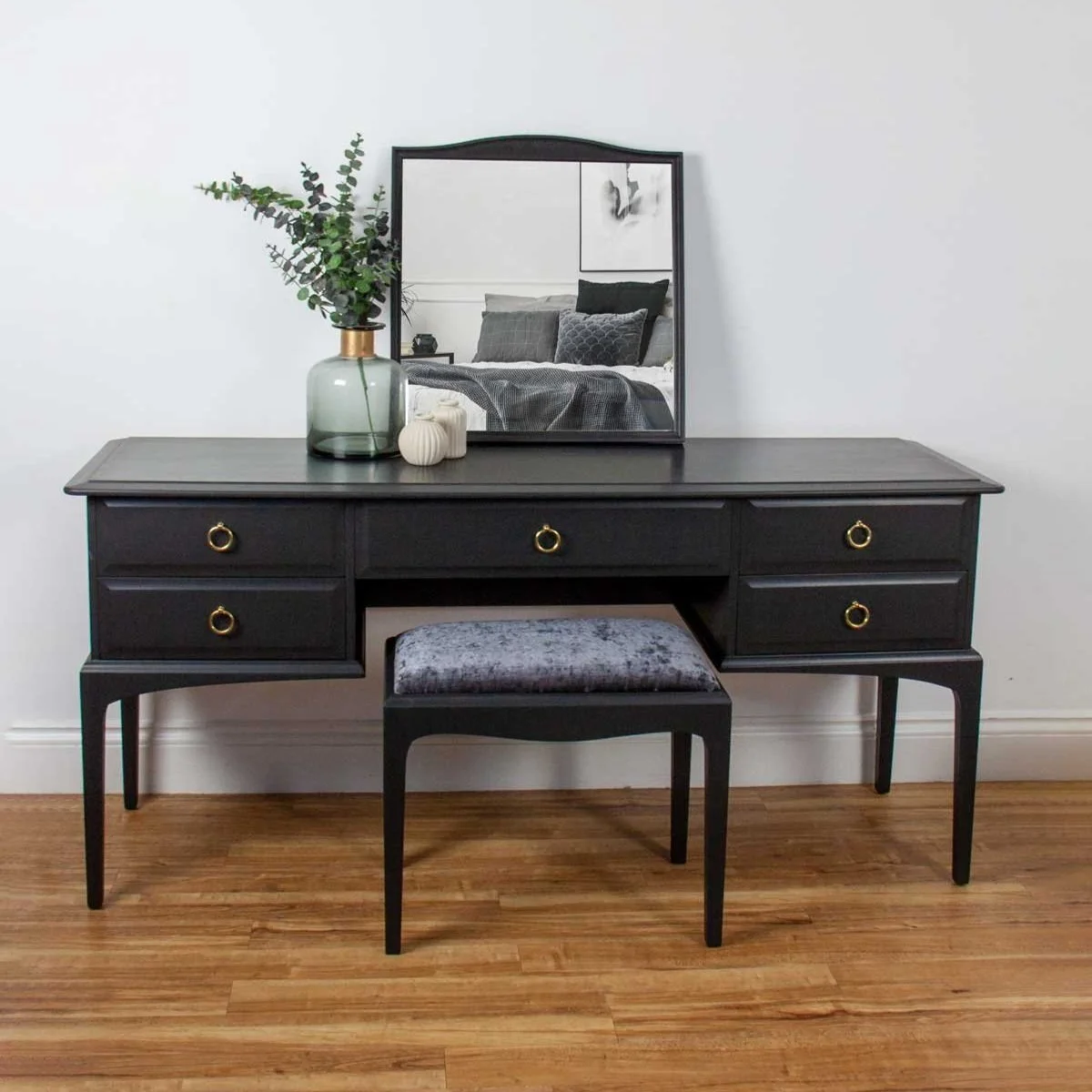 Black dresser with a mirror, a vase with greenery, and small decorative objects on top, set against a white wall with a bed reflected in the mirror.
