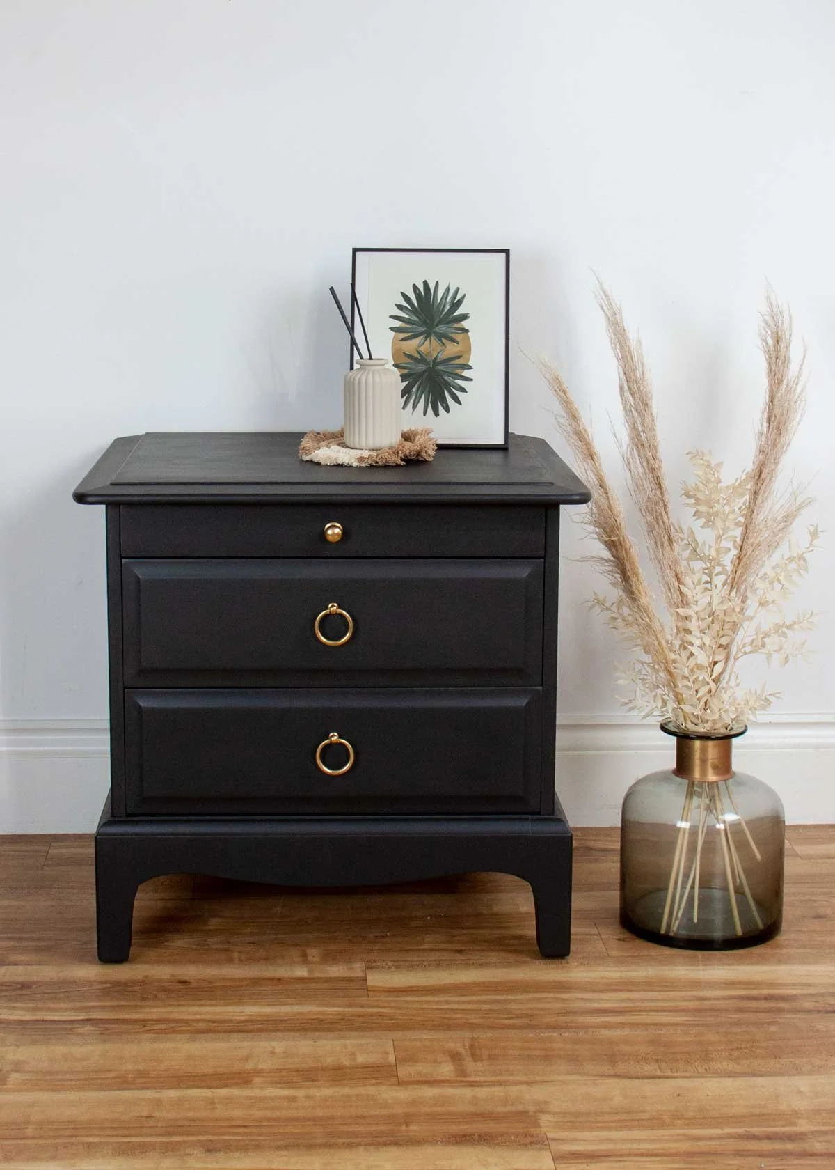 A black nightstand with three drawers, each with gold ring handles, placed against a white wall on a wooden floor. On top of the nightstand are a framed botanical print, a beige vase with black sticks, and a small decorative mat. To the right, there 