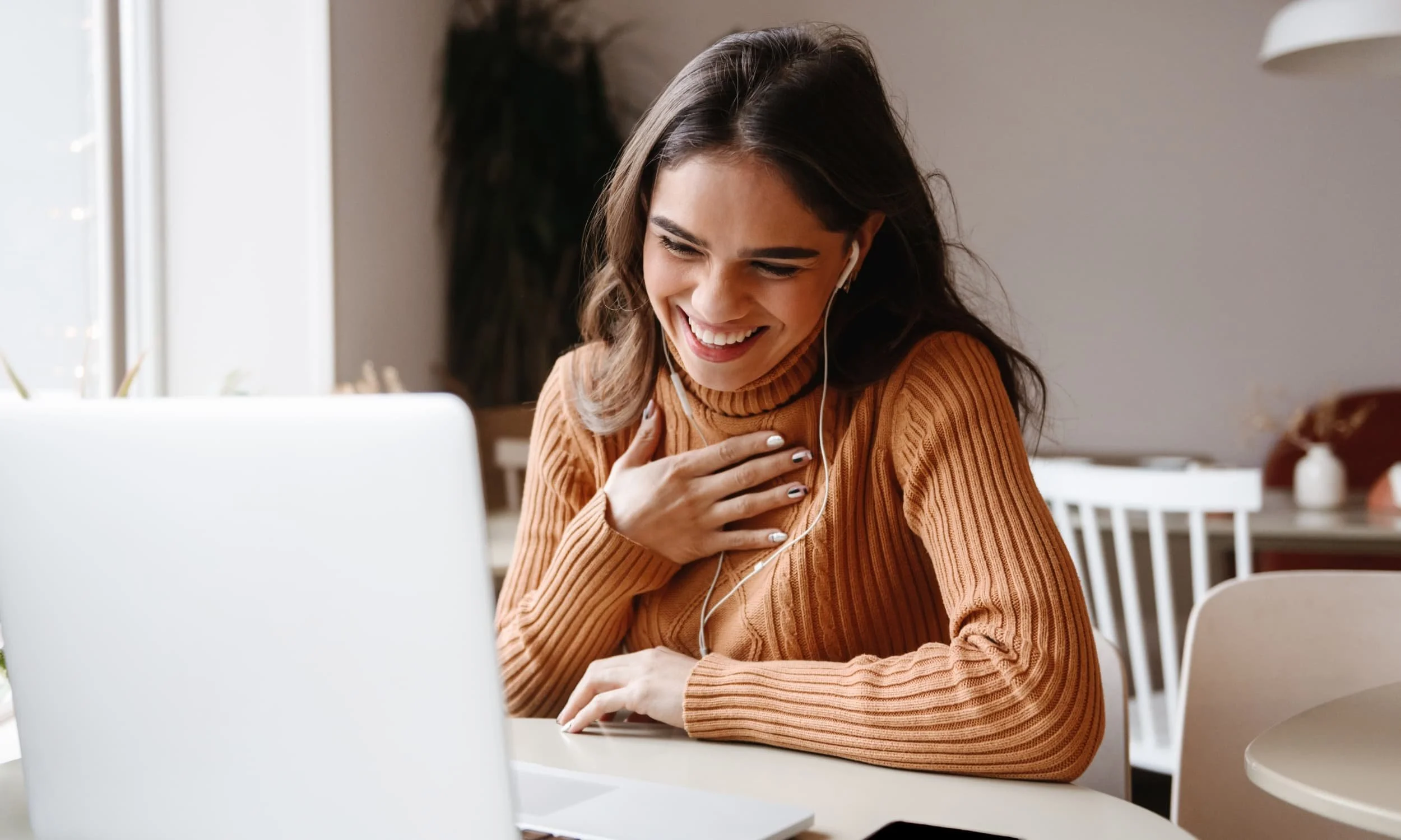 Lachende Frau mit beigem Pullover am Notebook mit Kopfhörern in den Ohren