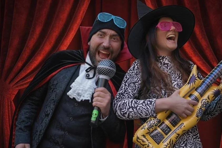 Two adults dressed in Halloween costumes performing a playful Lip Sync Dance Battle with props, including an inflatable guitar and microphone, during a Halloween event in London.