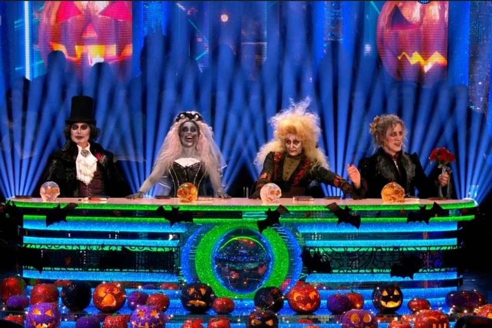 Four performers dressed in spooky Halloween costumes sitting behind a brightly lit judging panel on a Strictly-style stage, surrounded by glowing pumpkins and dramatic lighting.