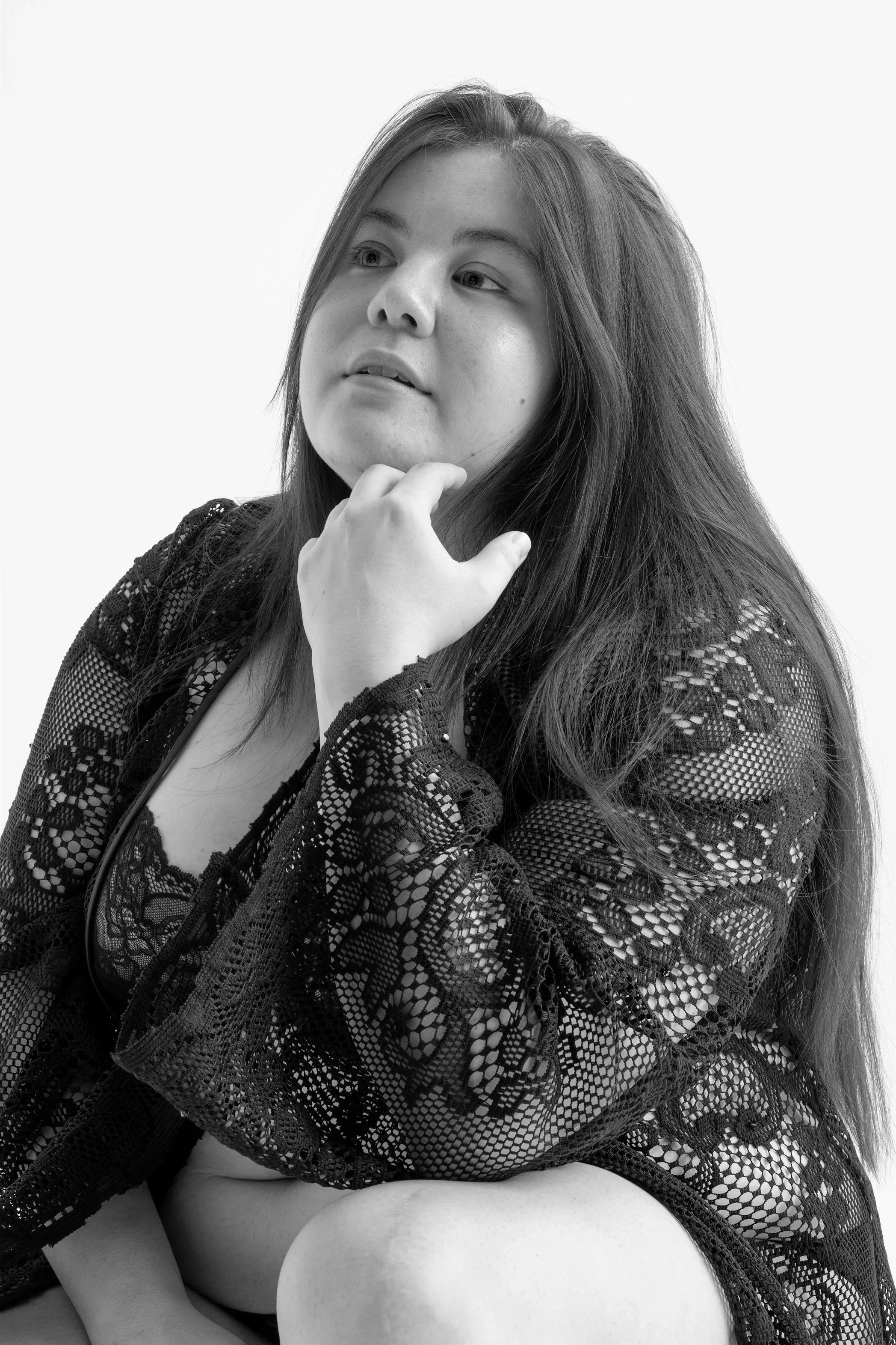 Portrait en noir et blanc d'une femme avec de longs cheveux, portant une chemise en dentelle, assise de côté, contemplant vers le haut, avec un fond blanc.