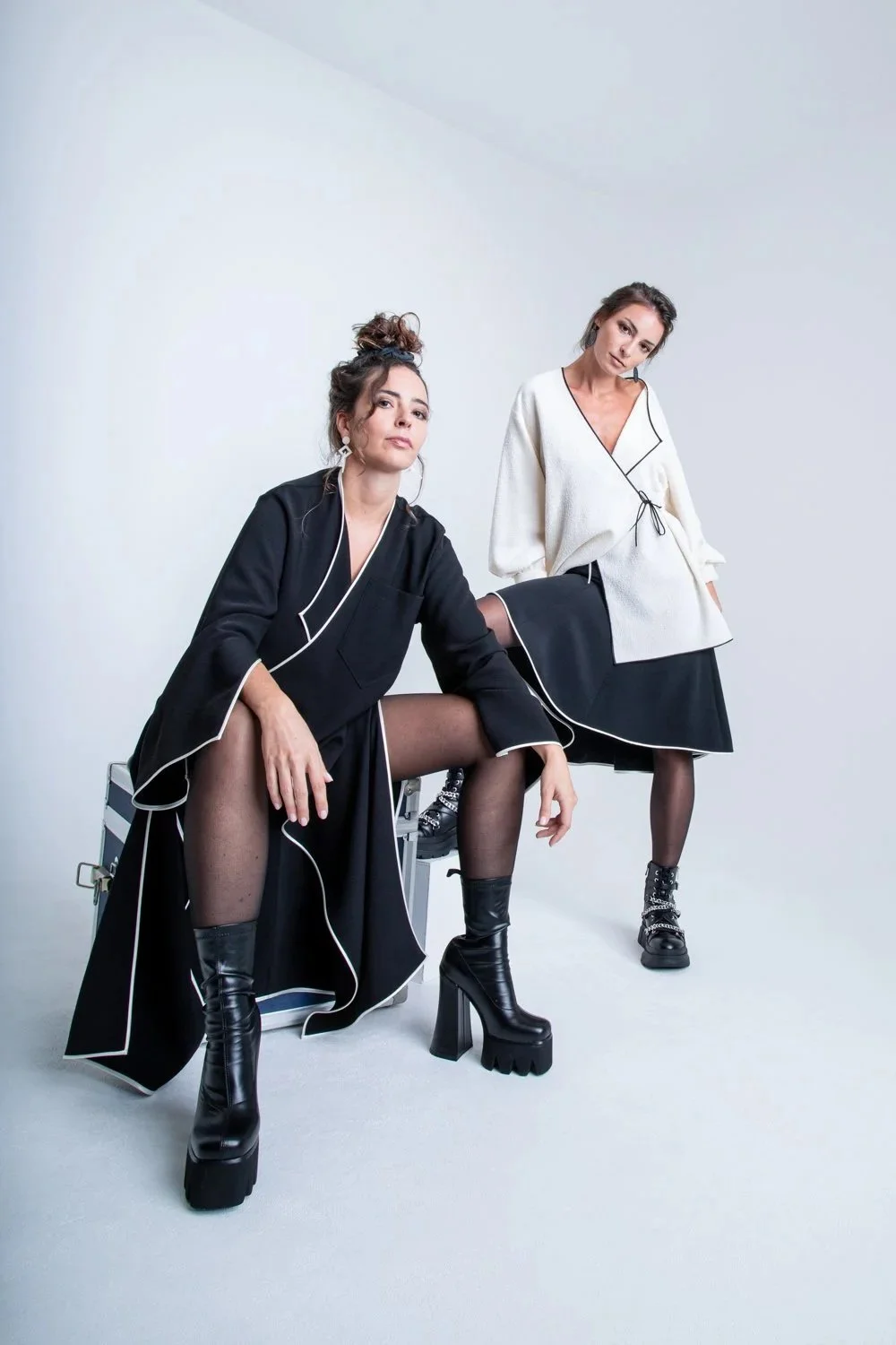 Deux femmes posant dans un studio blanc, portant des vêtements noirs et blancs avec des bottes à talons chunky, style moderne et fashion.