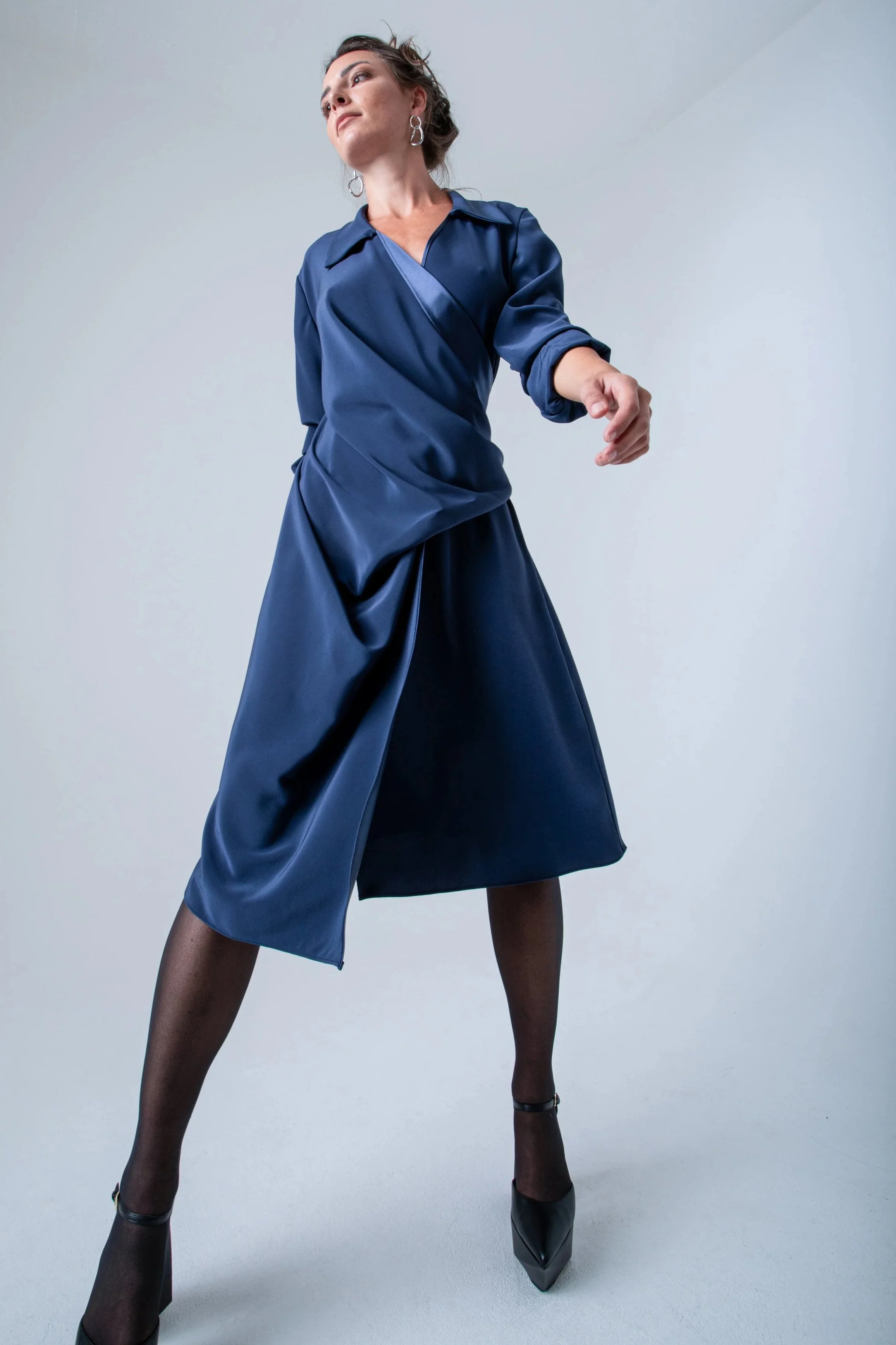 Une femme pose de manière confiante dans un studio contre un fond blanc, portant une tenue en satin bleu foncé avec des accessoires en argent, des collants noirs, des chaussures à talons noirs, et des boucles d'oreilles en forme de cercle.