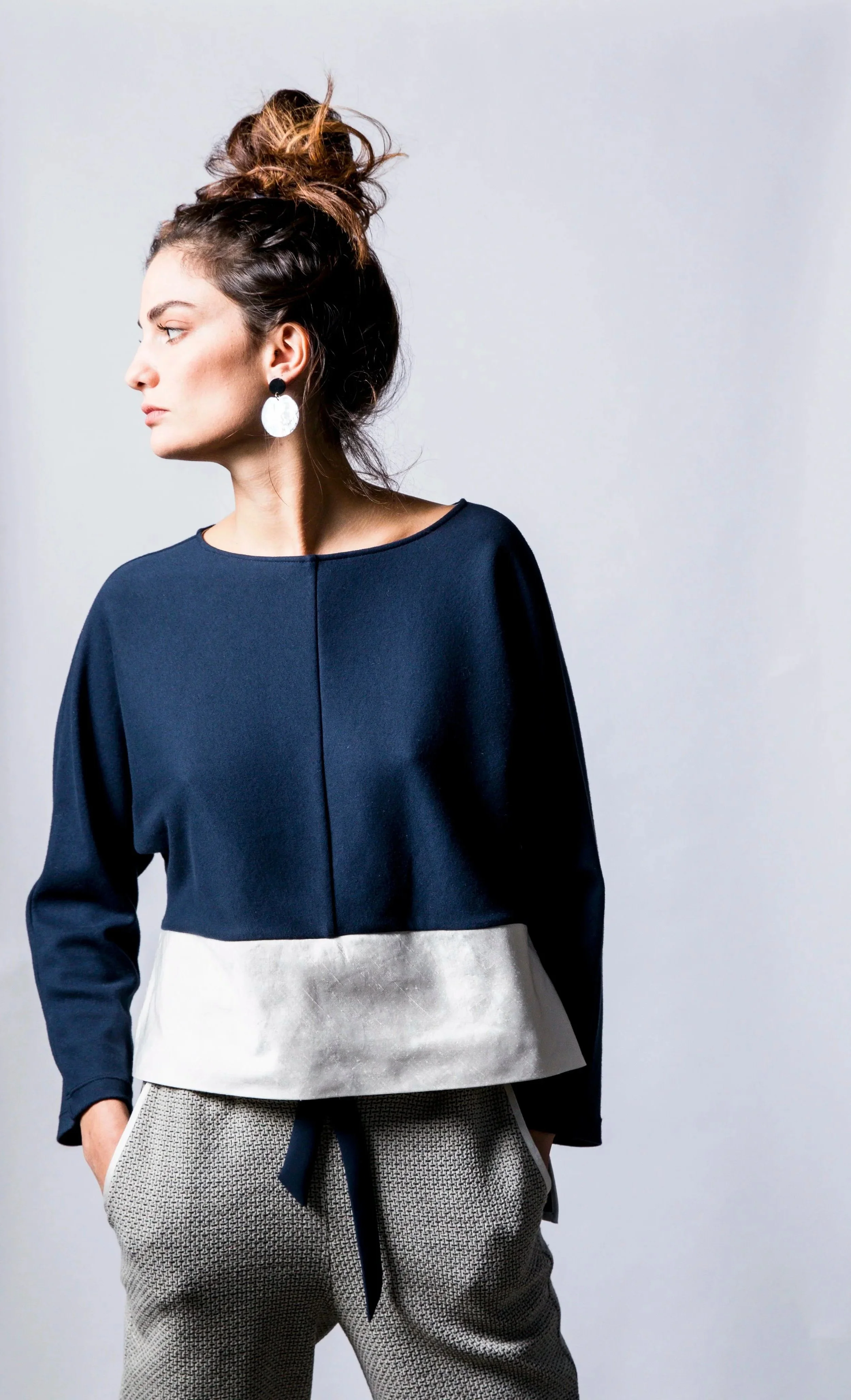 Une femme portant un haut bleu foncé avec une jupe beige à la taille et un pantalon à motifs gris et blanc, avec des boucles d'oreilles. Elle a les cheveux en chignon et regarde vers la gauche.