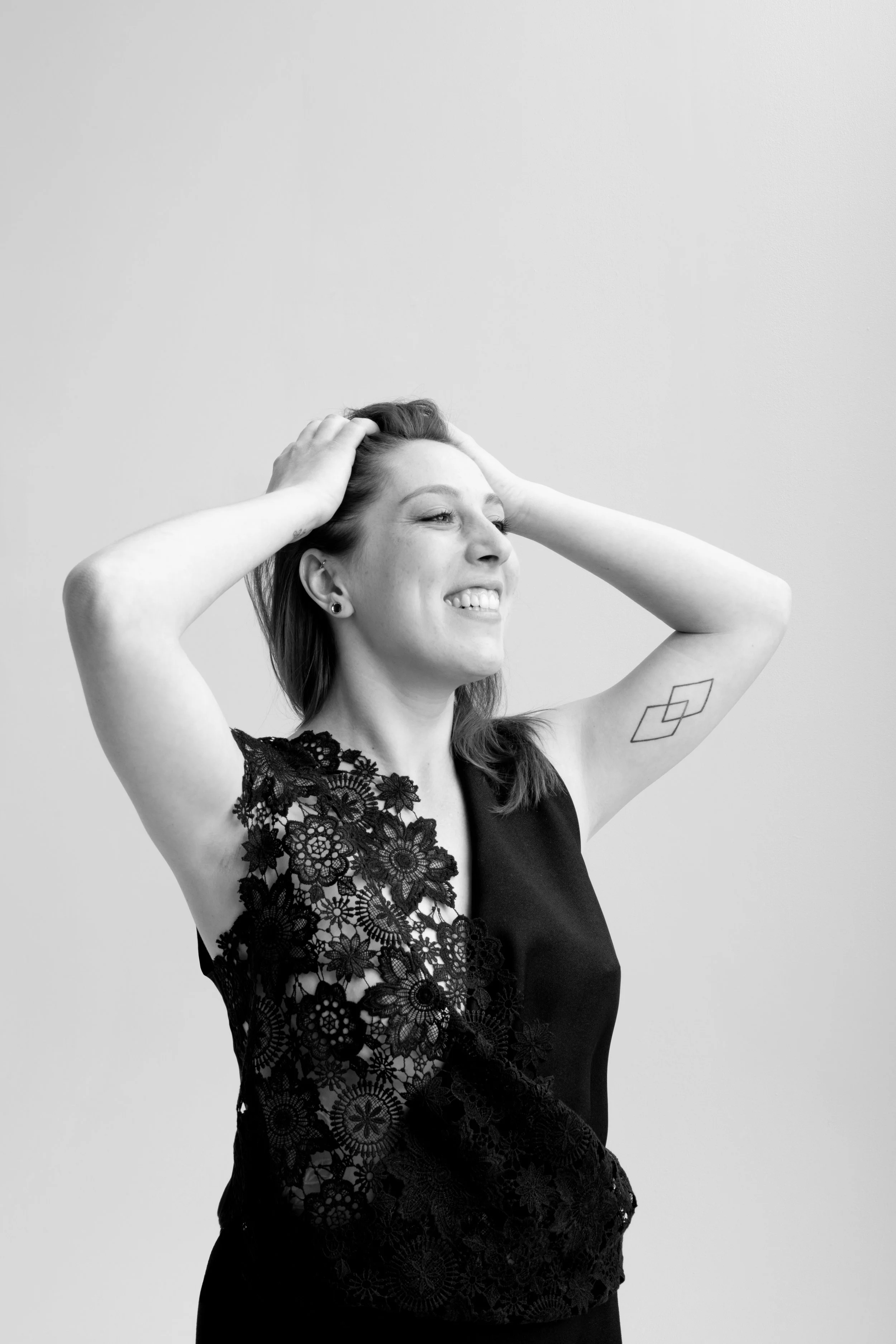 Une femme souriante en noir et blanc, tenant ses cheveux, portant une robe en dentelle avec un motif floral sur le côté gauche, posant à côté d'un mur blanc.