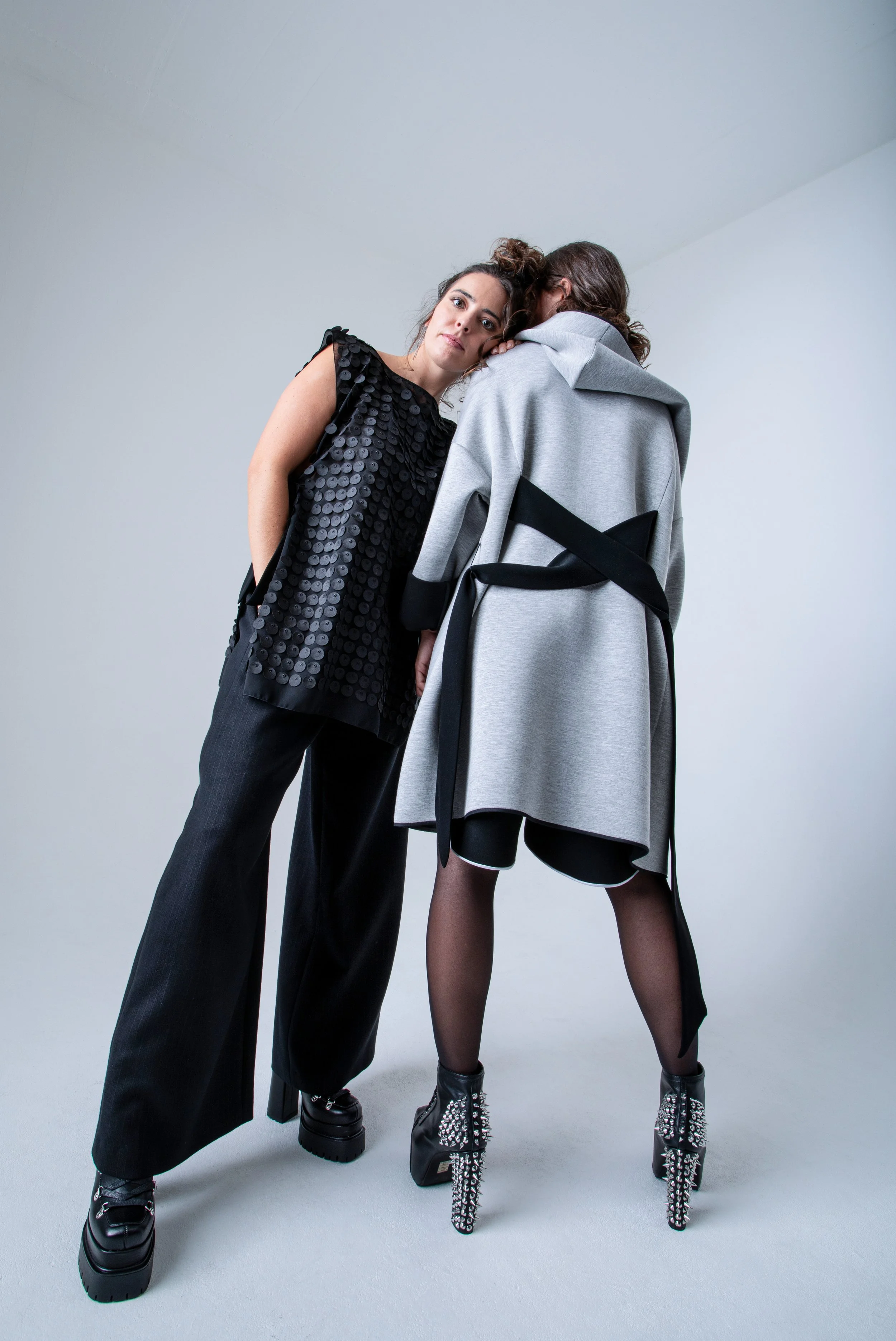 Deux femmes en vêtements de mode modernes posent sur un fond gris clair. L'une porte un haut noir à sequins et un pantalon noir, l'autre porte un manteau gris avec un design noir et des bottes à talons avec des détails en studs.