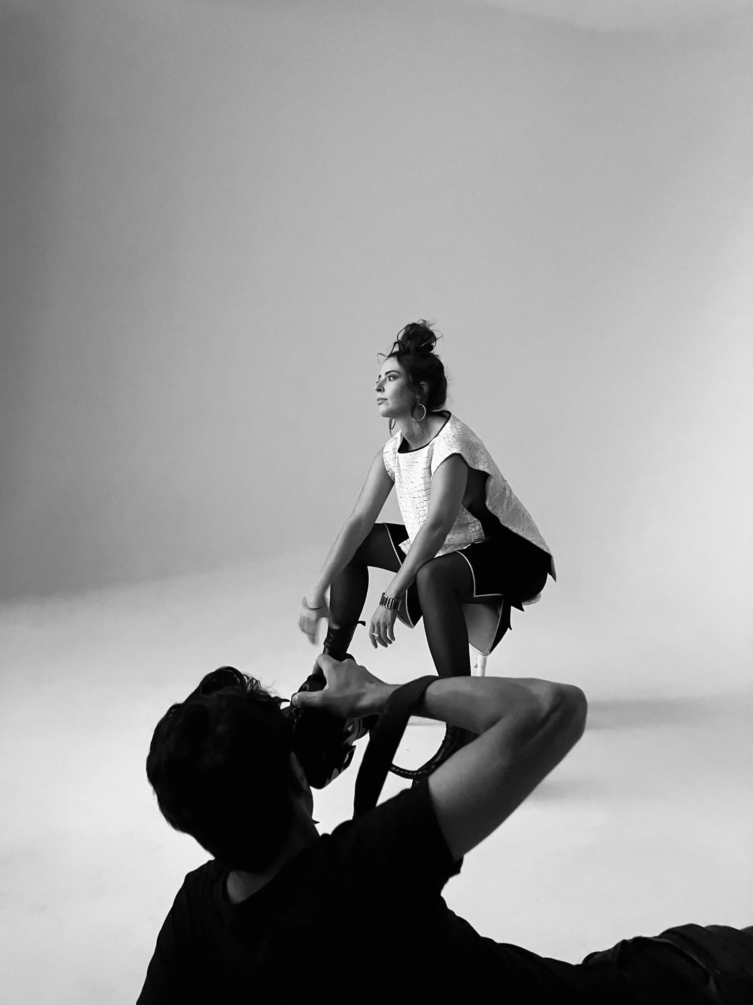 Une femme avec des cheveux attachés, portant une veste blanche et un short noir, assise sur un siège en studio avec un fond neutre. Une personne en premier plan, en position de prise de vue, semble aider la femme avec des équipements de tournage ou d