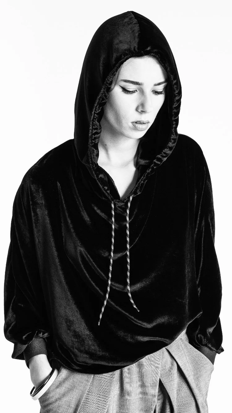 Une femme porte un sweat à capuche noir en velours avec un col en V, elle a les yeux baissés et les mains dans les poches d'un pantalon à carreaux.