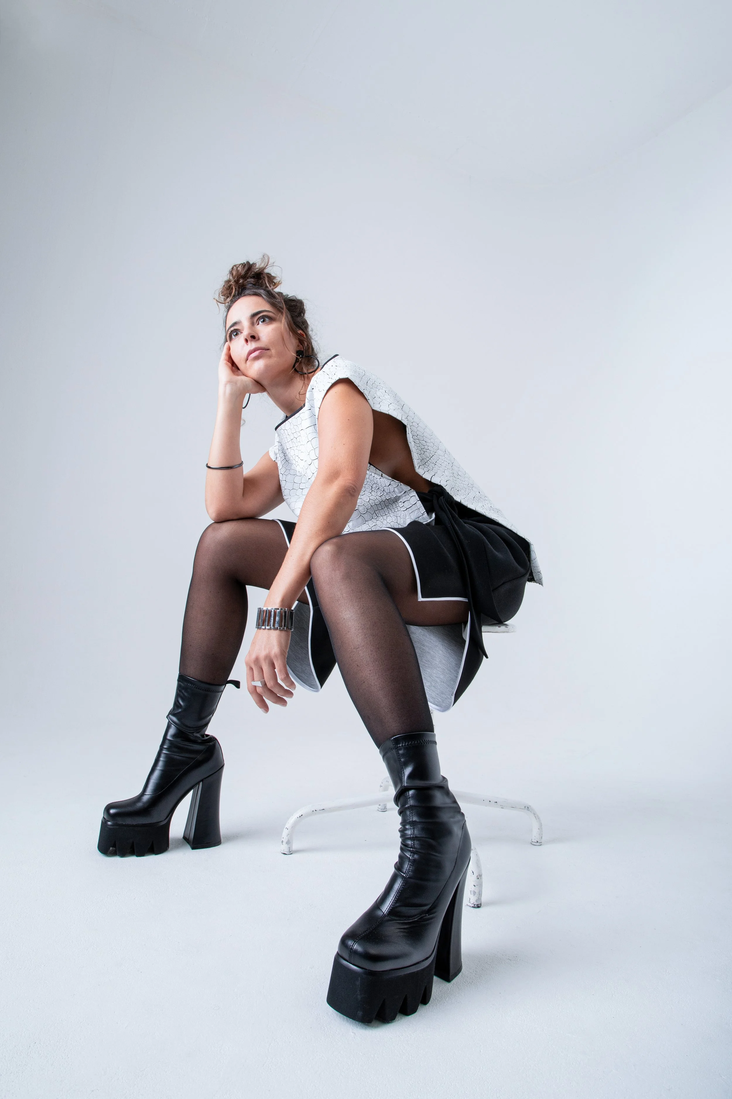 Une femme assise sur une chaise blanche, portant une tenue noire et blanche avec des chaussures à talons très épaisses et à plateforme, en pose décontractée et pensive dans un fond neutre.
