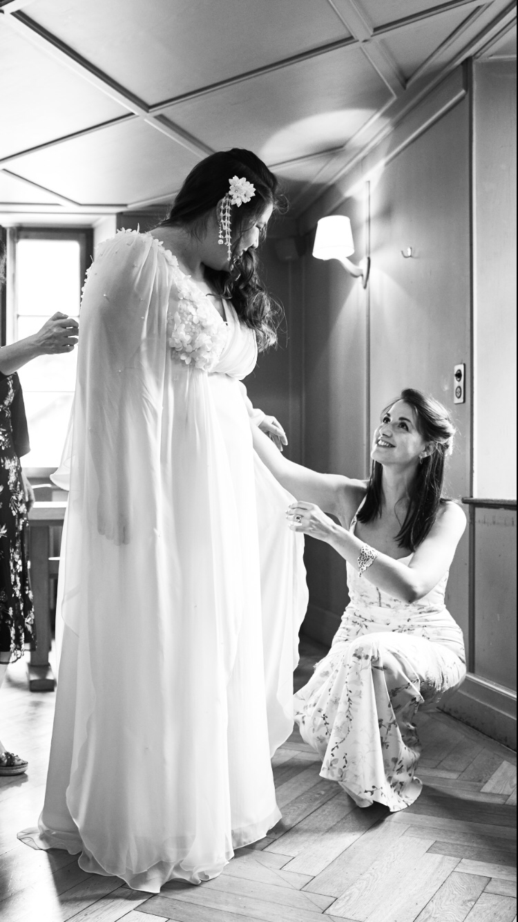 Une femme habillée en robe de mariée est aidée à ajuster sa robe par une autre femme, dans une pièce éclairée par une lampe murale, en noir et blanc.