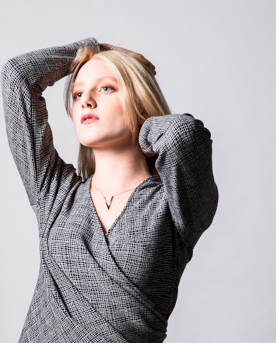 Femme blonde posant avec bras levés, portant une veste à motifs et un collier