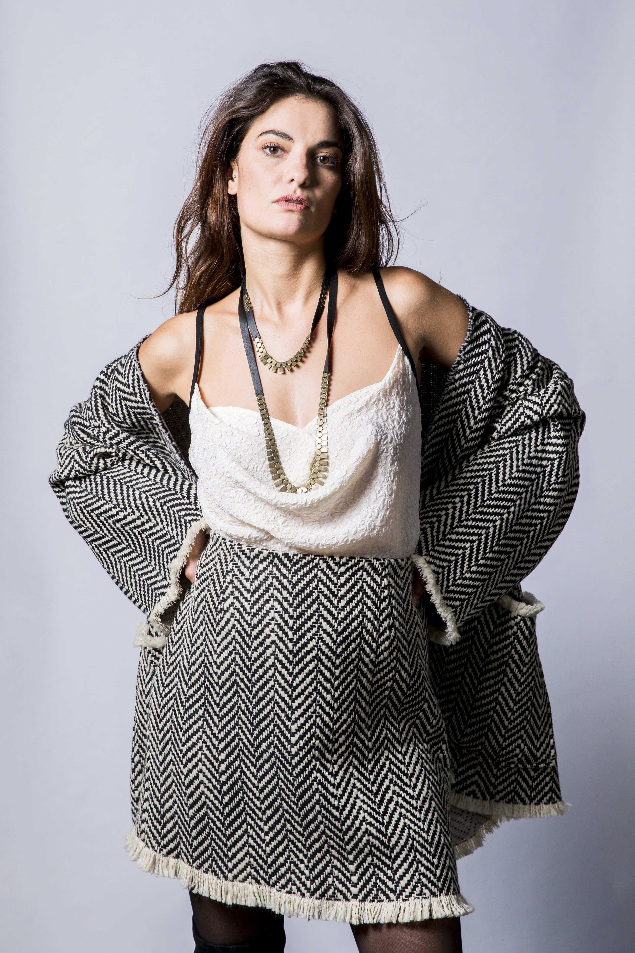 Une femme avec des cheveux bruns longs, portant un collier doré, une robe blanche en tissu texturé, une veste ou un pull en laine à motif zigzag noir et blanc, et des collants noirs, pose contre un fond gris clair.
