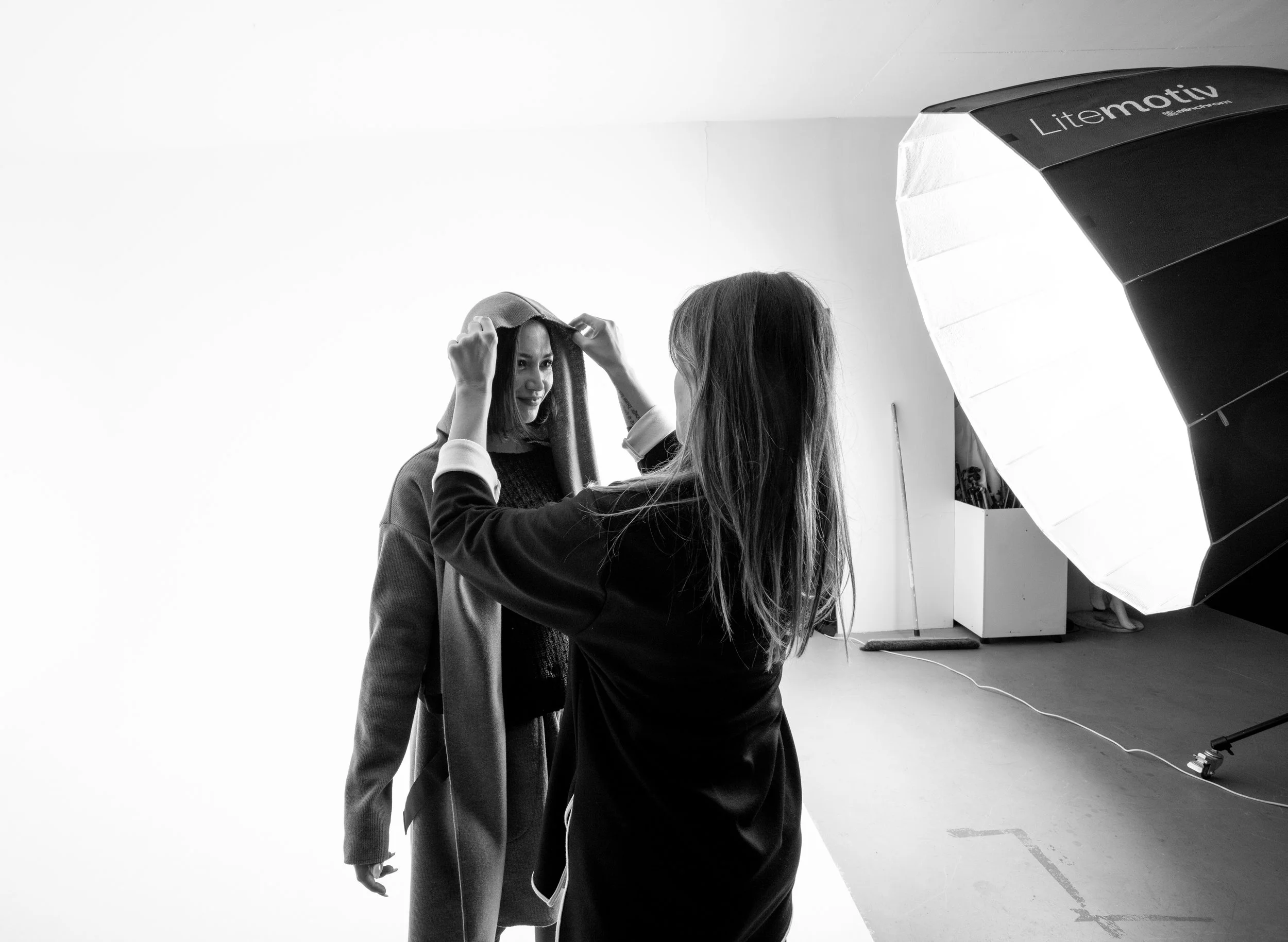 Deux femmes dans un studio photo, l'une ajuste un manteau sur l'autre, avec un flash de studio et un fond blanc.