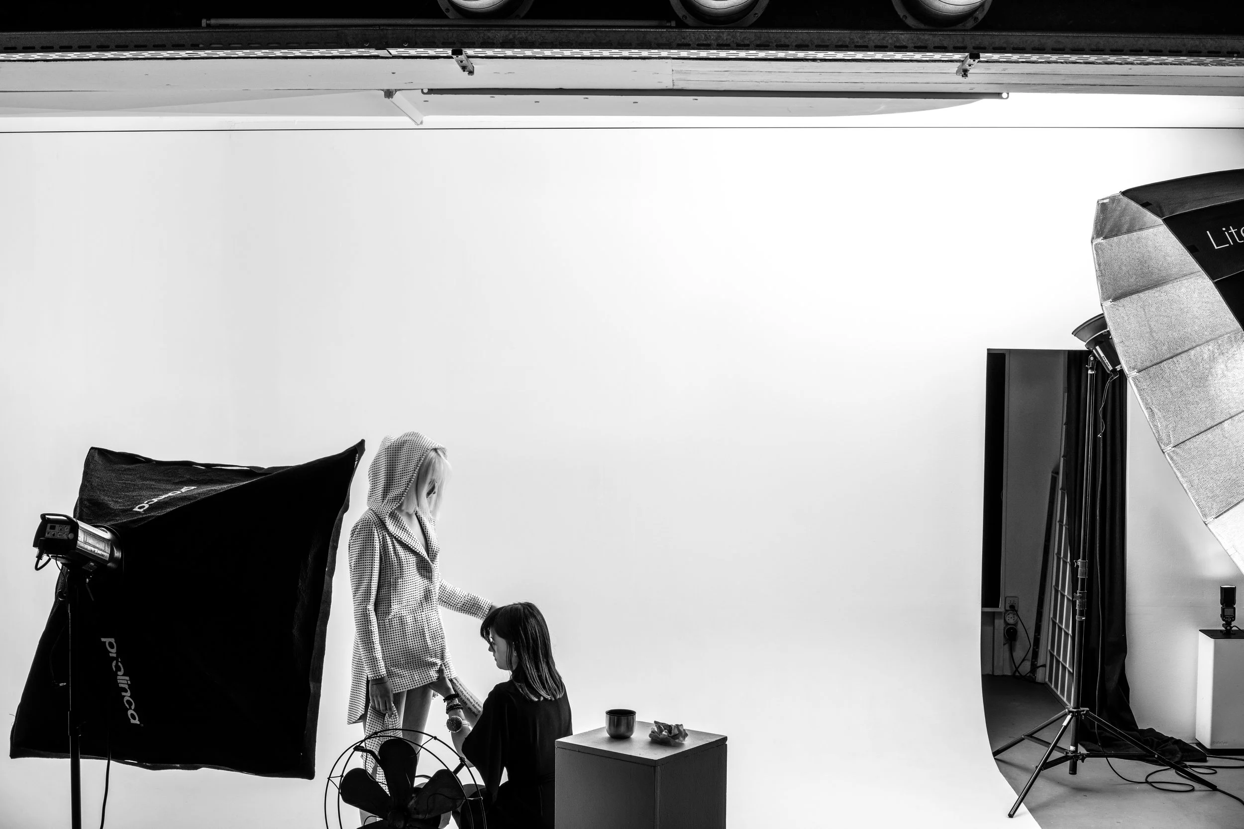 Une séance photo en studio avec un fond blanc, éclairages et équipement de photographie. Deux jeunes filles debout discutent, une d'elles a une robe à pois, l'autre porte des vêtements décontractés. Il y a un parapluie lumineux, un ordinateur portable, une table avec un mug, et des accessoires de studio.