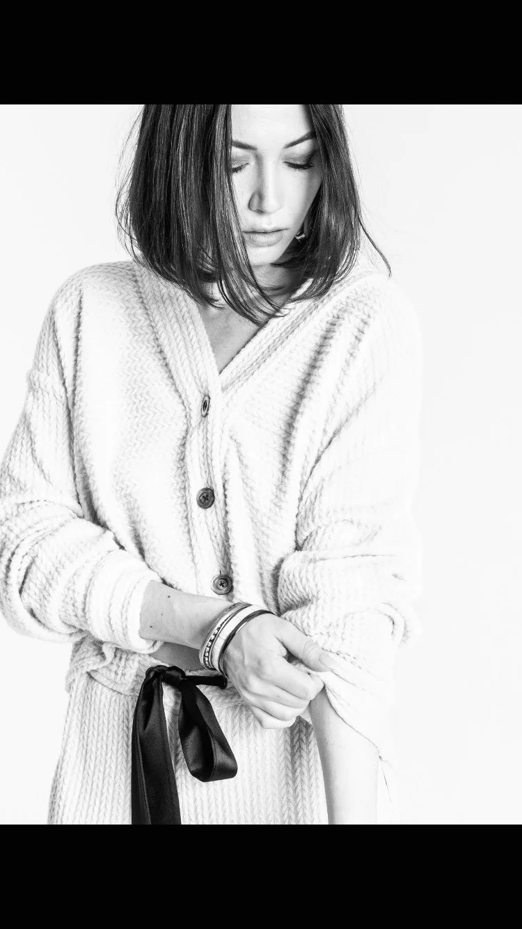 Une femme avec des cheveux courts et bouclés, portant un pull en laine clair avec des boutons, ajuste le bas de son manche. Elle porte plusieurs bracelets à son poignet gauche et est en noir et blanc.