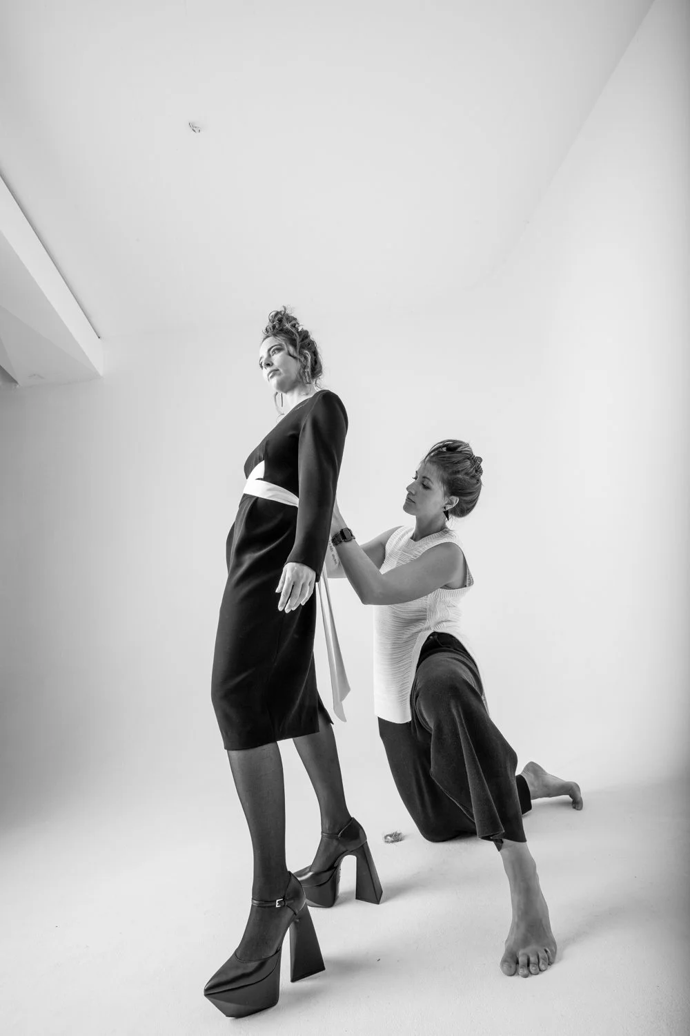 Deux femmes dans une session de couture ou de maquillage, l'une debout en robe noire et talons hauts, l'autre à genoux ajustant la robe, dans un environnement minimaliste et lumineux.