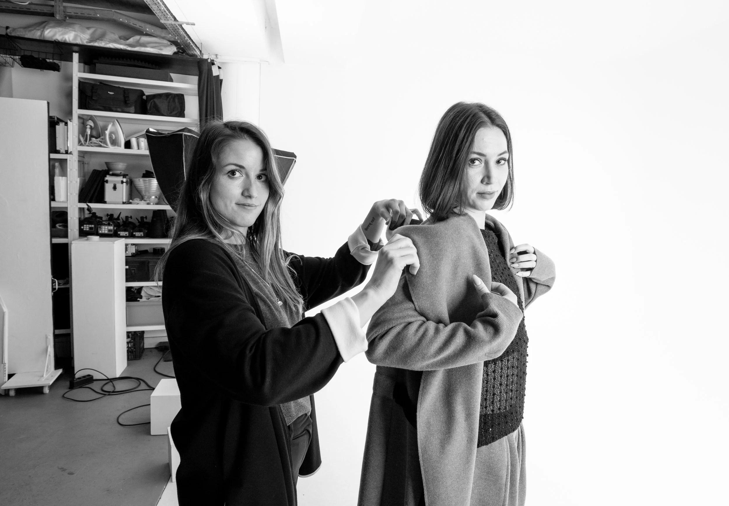 Deux jeunes femmes dans un studio, l'une ajustant le col de l'autre, qui porte un manteau ou une veste. En arrière-plan, des étagères avec des objets divers, y compris des appareils électroniques et des boîtes.