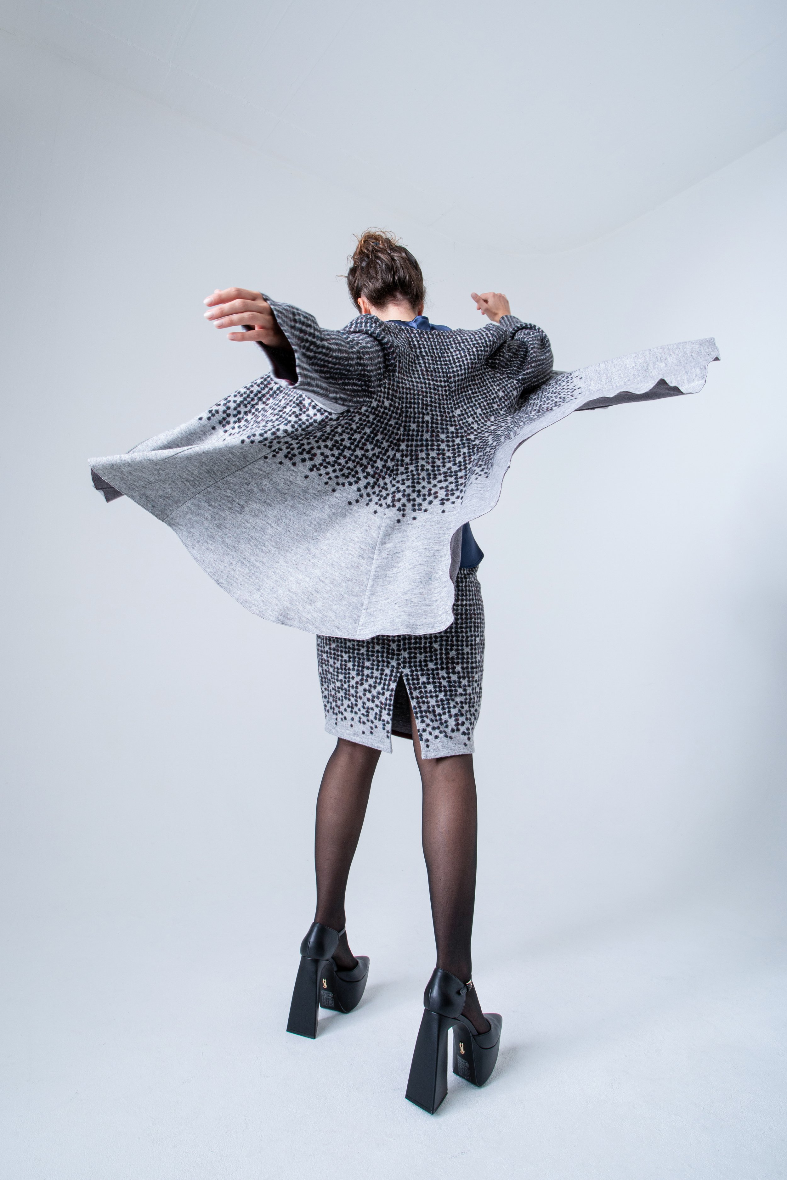 Une personne vue de dos, portant une veste à motifs noire et blanche, une jupe assortie, des collants noirs, des chaussures à talons épaules épaisses, et une paire de bottes à plateforme.