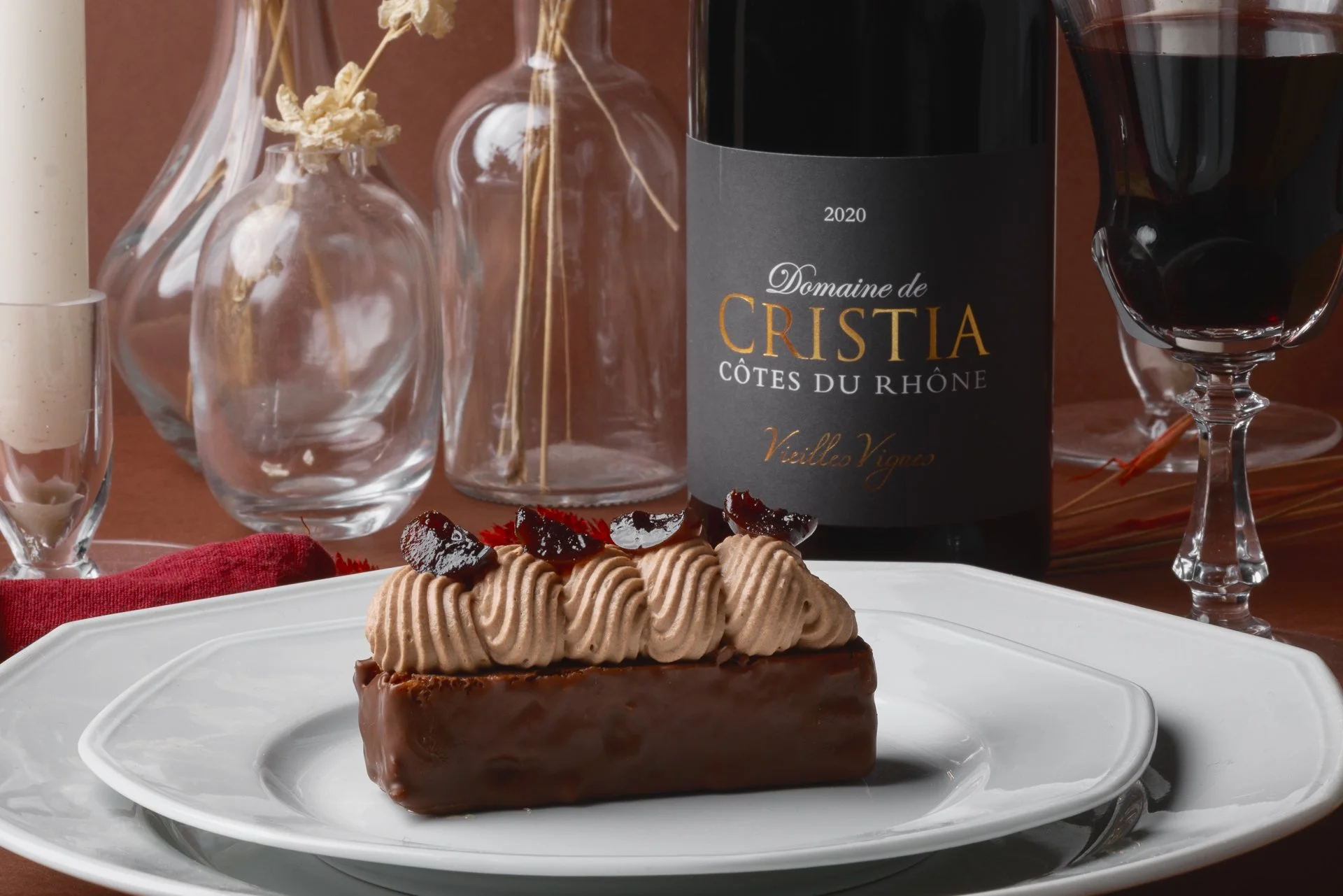 Gâteau au chocolat avec une décoration de crème et de morceaux de cerise, sur une assiette blanche. En arrière-plan, une bouteille de vin rouge, un verre de vin, et des vases en verre avec des branches séchées.