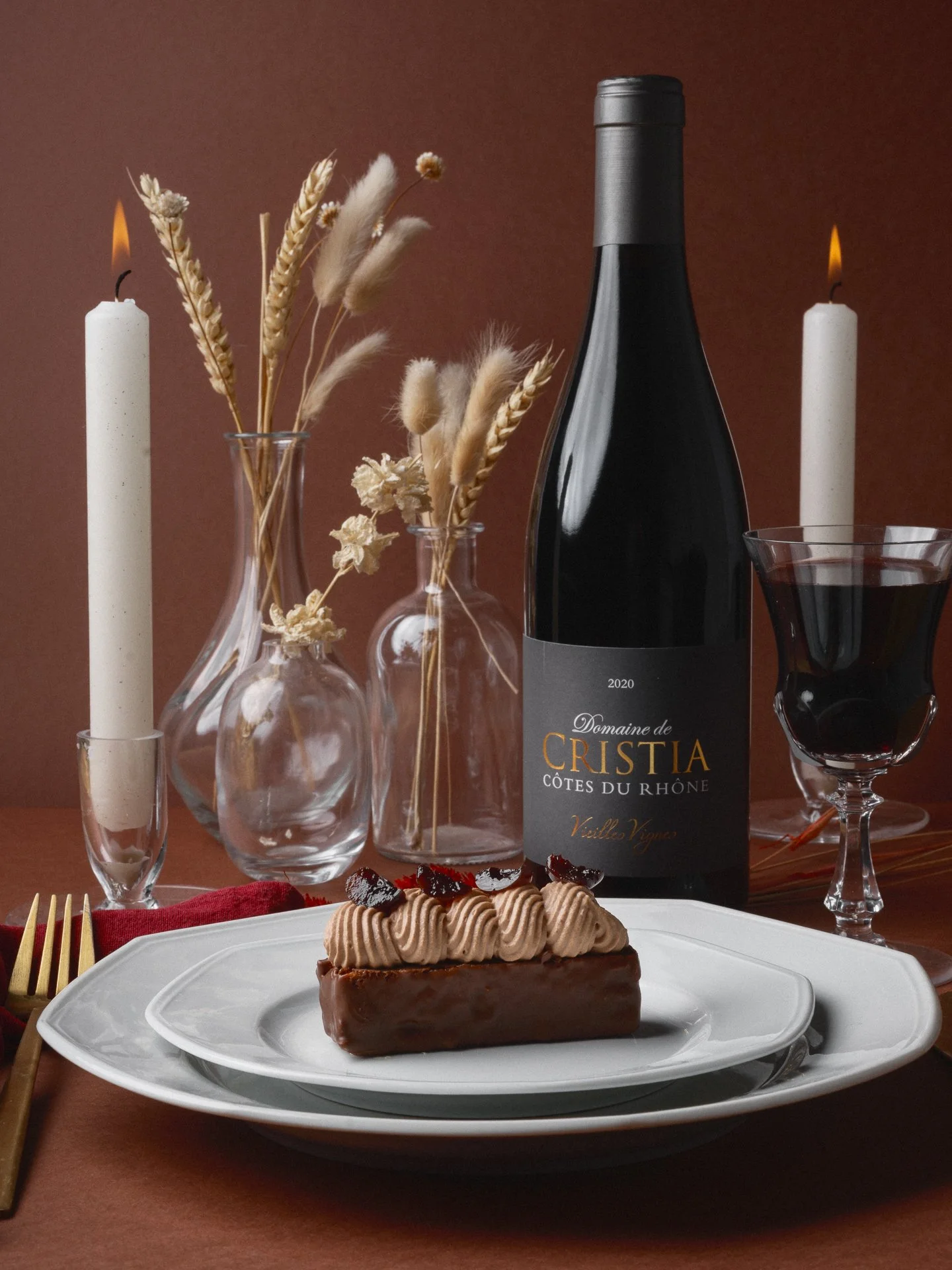 Une assiette de dessert au chocolat avec une décoration de confiture ou de gelée, accompagnée d'une bouteille de vin rouge, deux bougies allumées, des vases avec des fleurs sèches et un verre de vin rouge, avec une nappe rouge.
