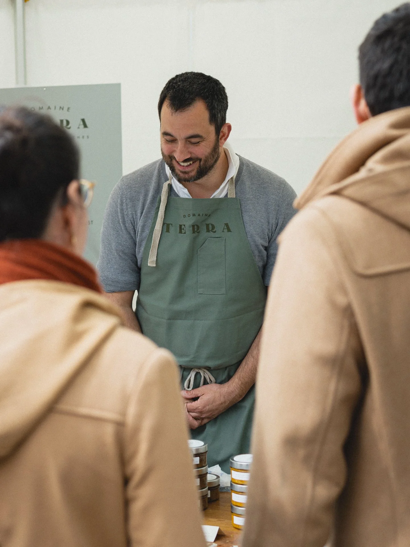 Un homme avec une barbe, portant un tablier vert, vend des produits en pot lors d'une foire ou d'un marché, entouré d'autres personnes.