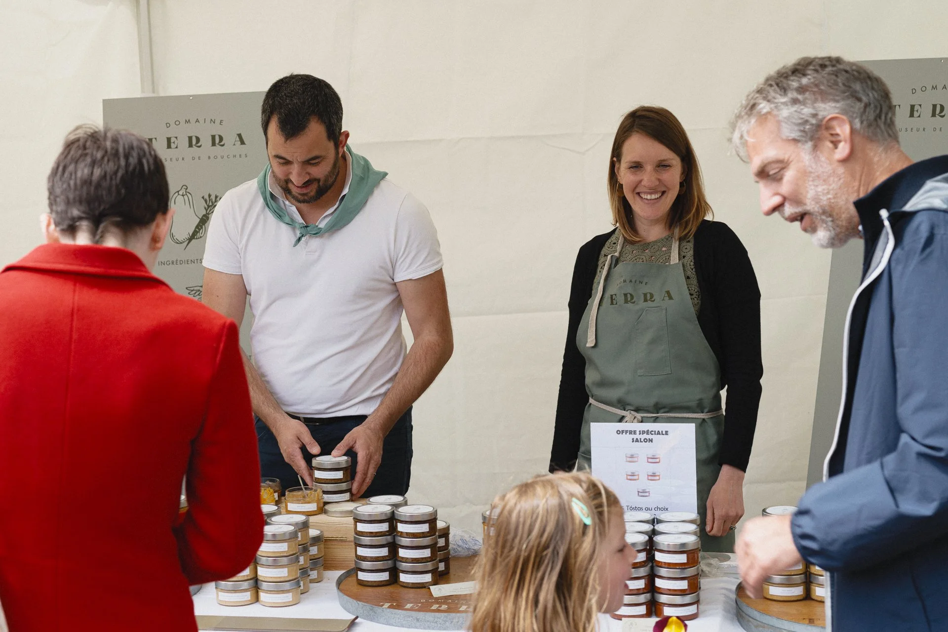 Groupe de personnes échangeant des produits en petits pots lors d'un événement. Une femme porte un tablier avec le logo 'Domaine Terra'.