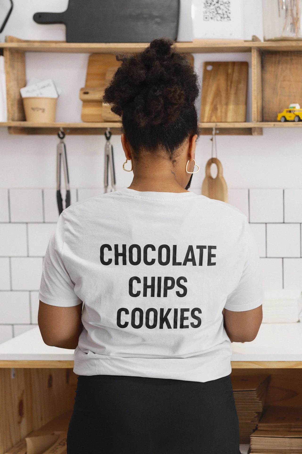 Une personne vue de dos portant un t-shirt blanc avec l'inscription « CHOCOLATE CHIPS COOKIES » en noir, dans une cuisine avec étagères en bois.