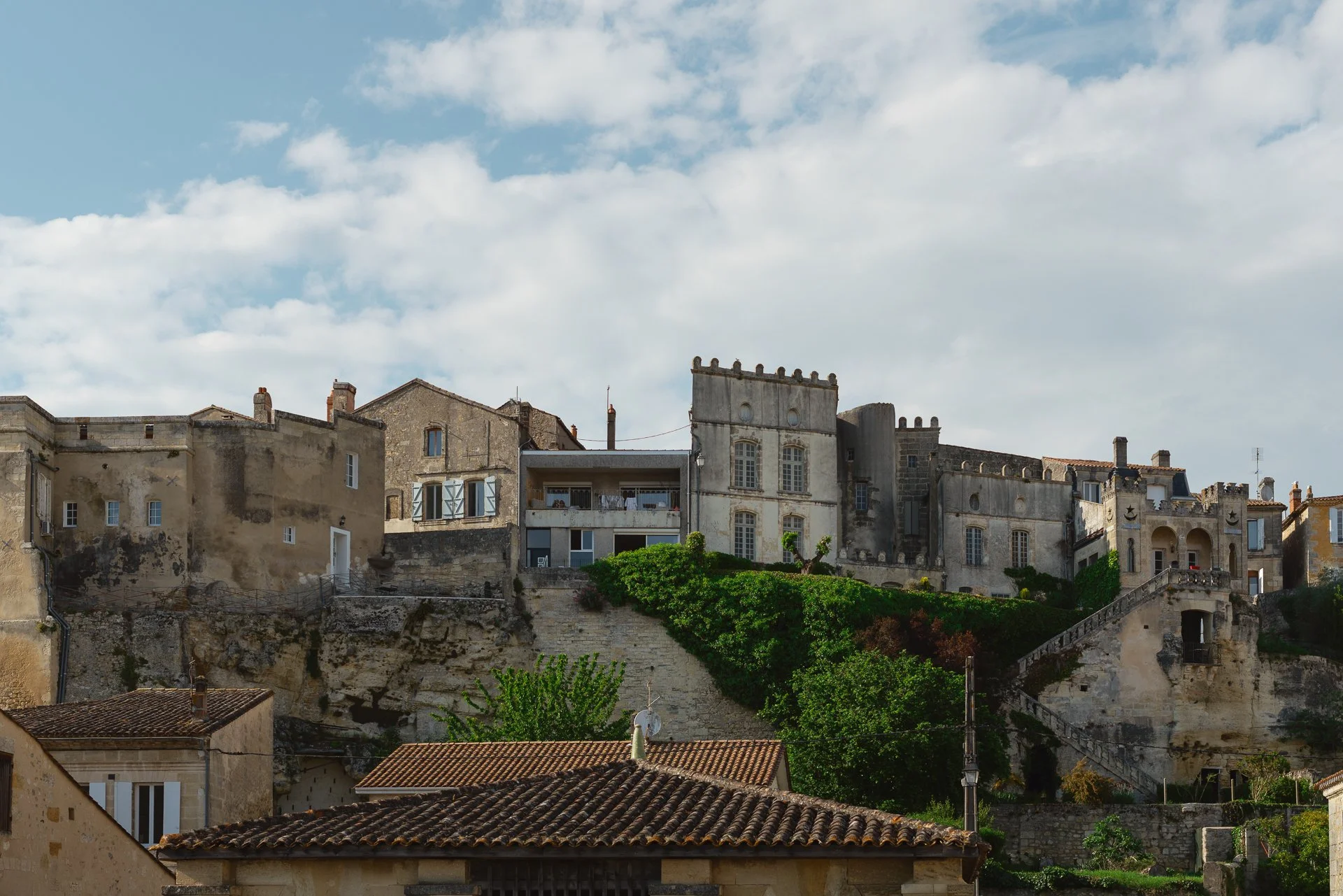 Villas anciennes en pierre sur une colline, avec escaliers en pierre et végétation verte, sous un ciel nuageux. Adapté au style de la ville de Saint-Paul-de-Vence, en France.