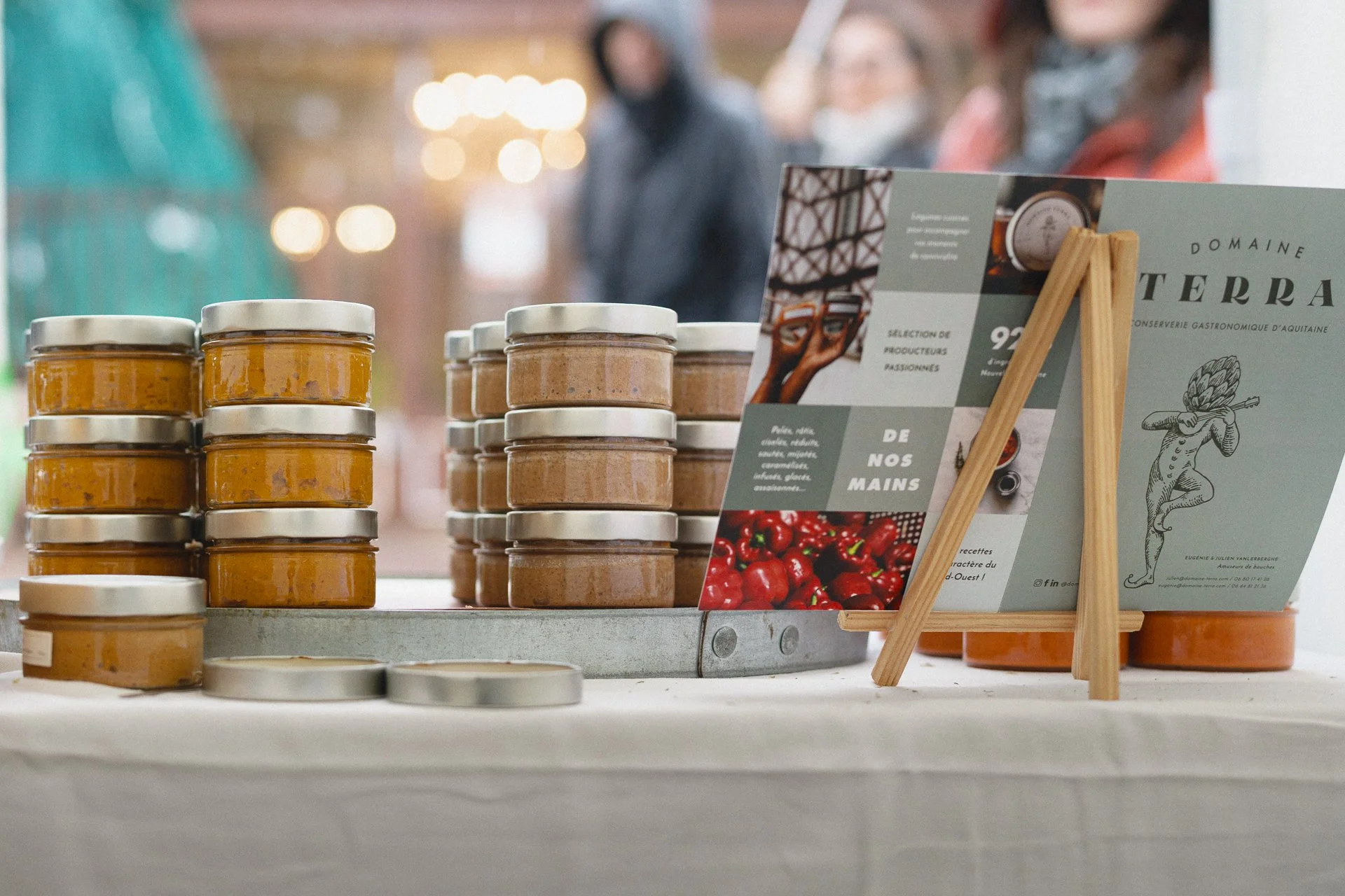 Pots de confiture ou de chutney empilés sur une table, avec un panneau d'information sur la dégustation et la production locale en arrière-plan.