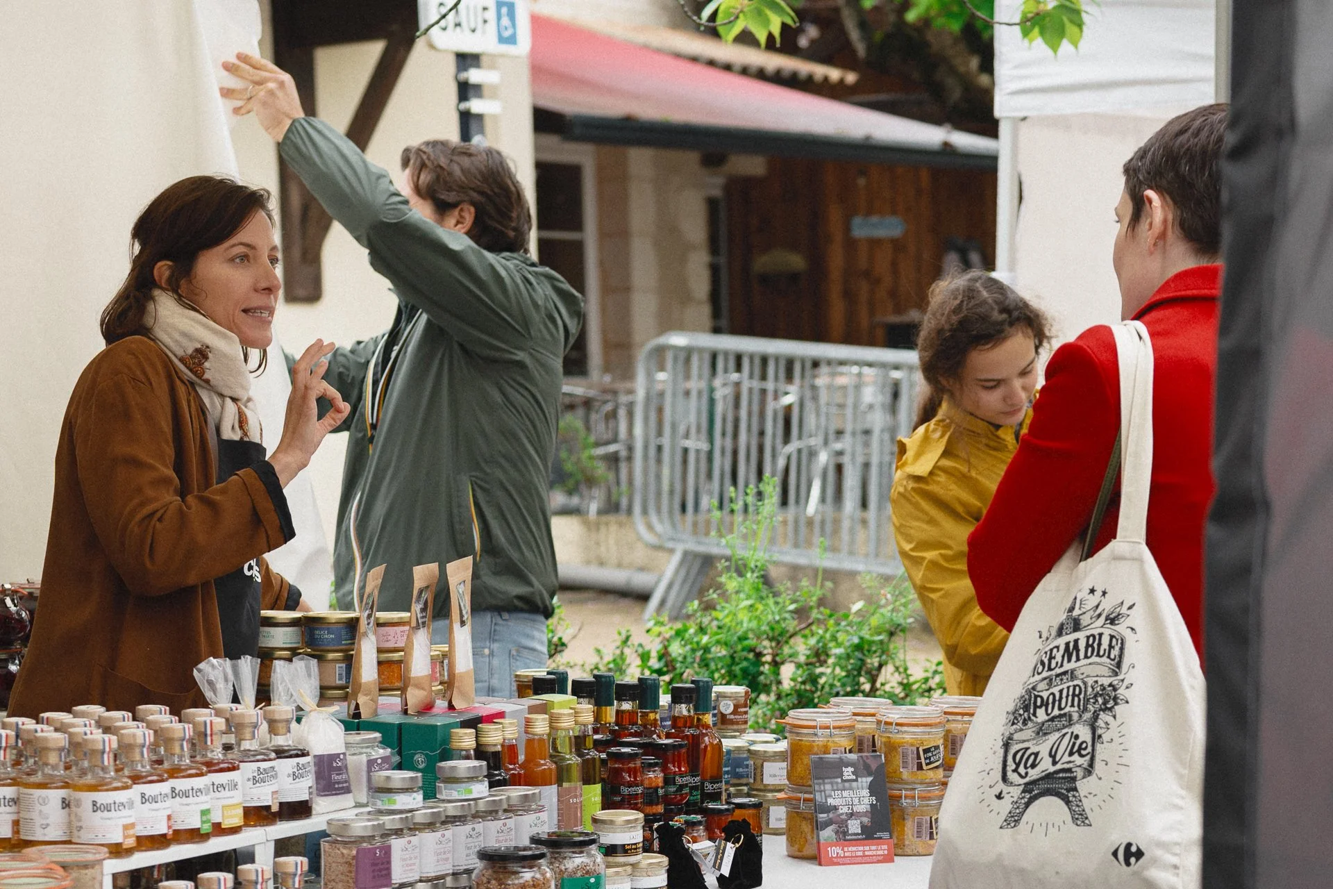 Une scène de marché en plein air avec plusieurs personnes se renseignant sur des produits. Un stand avec divers pots de confitures, huiles et autres aliments artisanaux est visible au premier plan. Une femme explique quelque chose à un homme tout en 