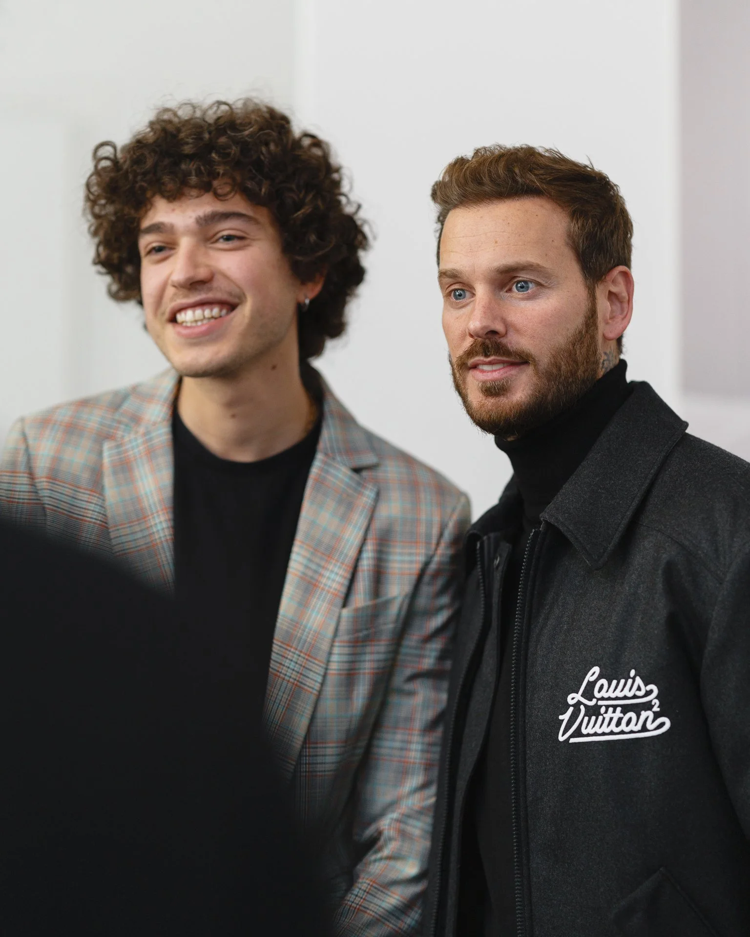 Deux hommes posant pour une photo, l'un porte une veste en tweed avec un t-shirt noir, l'autre porte une veste noire avec une capuche Louis Vuitton.