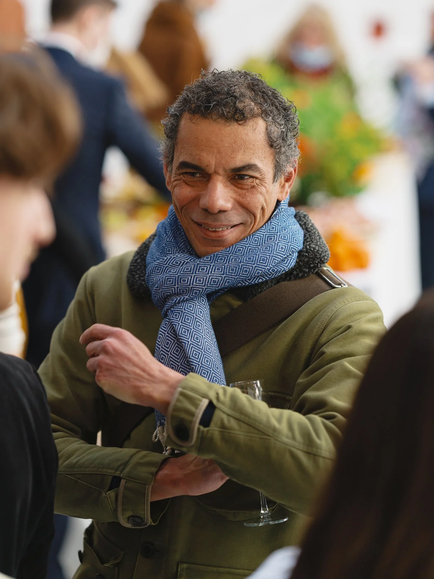 Un homme souriant lors d'une conversation dans un cadre social, avec plusieurs personnes floues en arrière-plan.