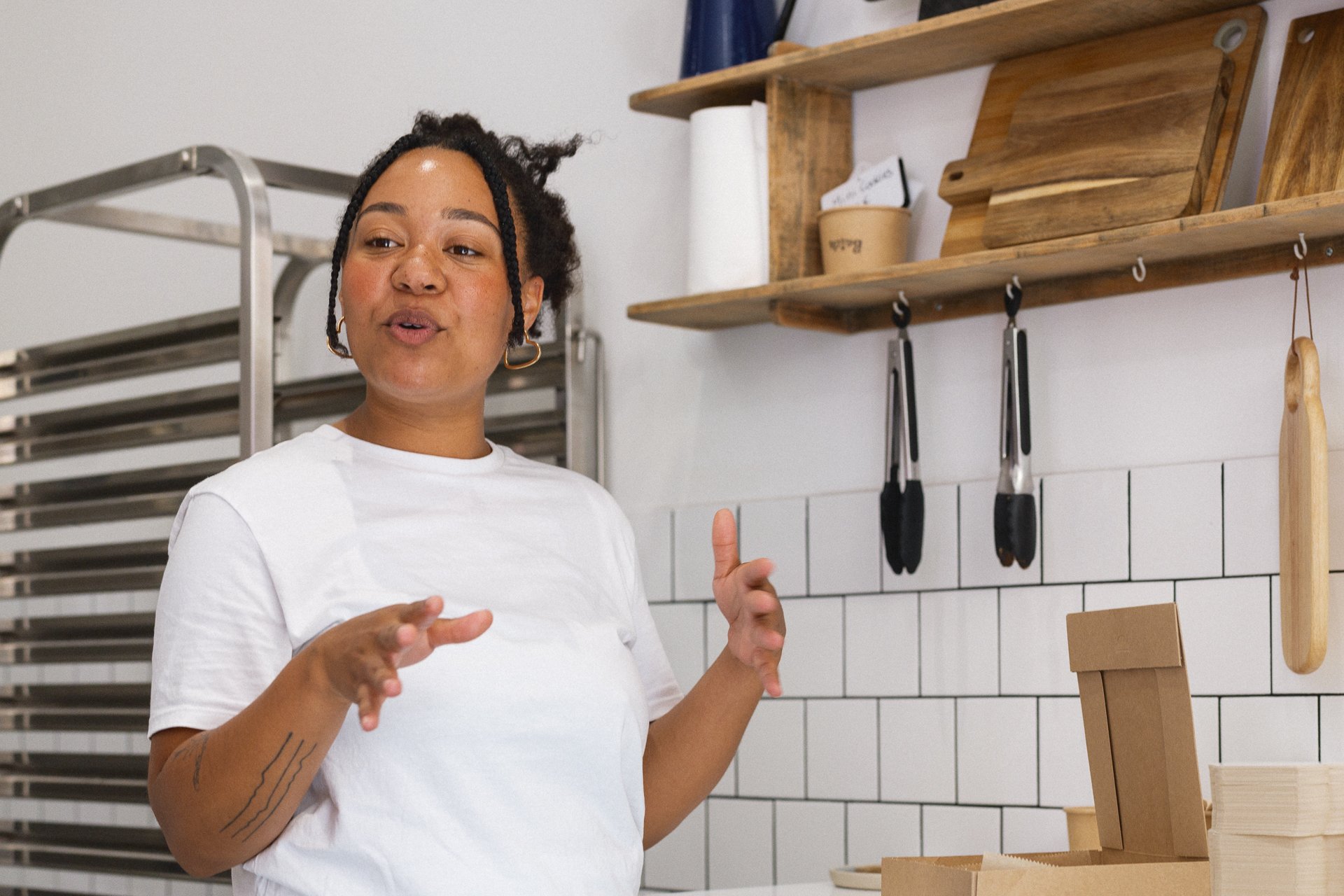 Une femme avec des cheveux bouclés courts, portant un t-shirt blanc, parle dans une cuisine ou un atelier avec des étagères en bois et des ustensiles suspendus.