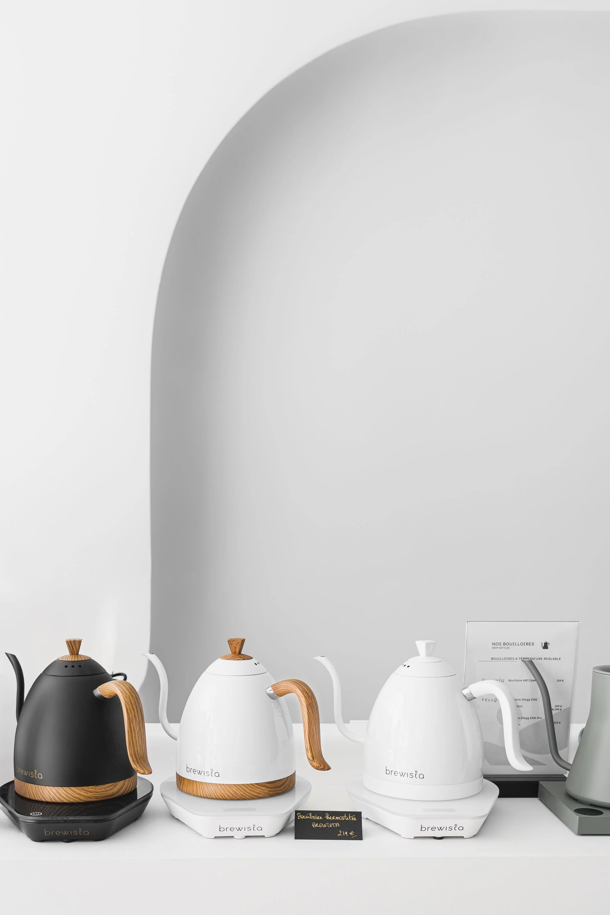 Une rangée de plusieurs bouilloires électriques de couleurs différentes sur un comptoir blanc, avec un mur blanc en arrière-plan. Une affiche noire avec une photo en noir et blanc et la marque SIBARIS.