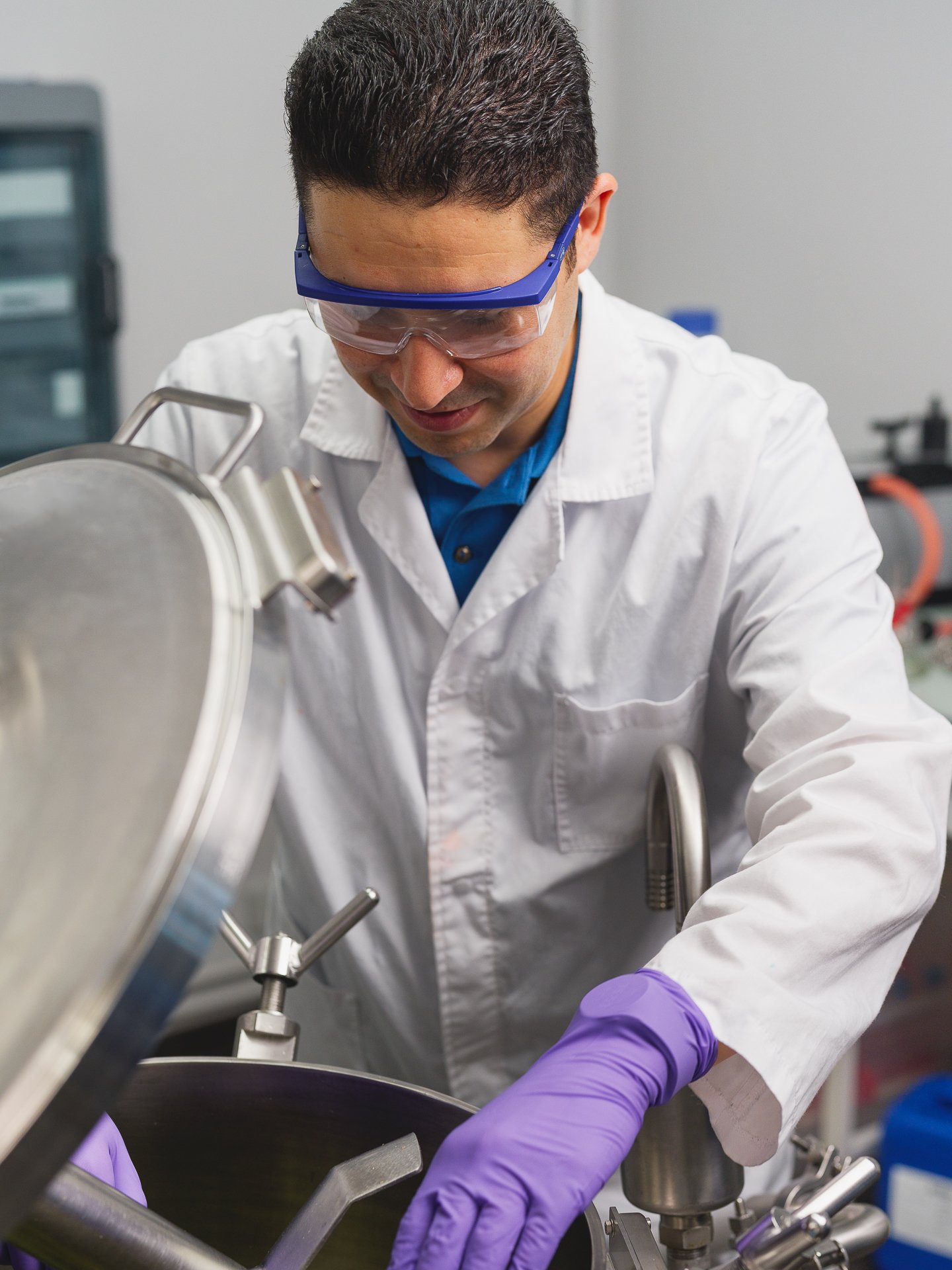 Un scientifique portant des lunettes de protection et un manteau blanc travaille avec un équipement en laboratoire, manipulant un objet avec des gants violets.