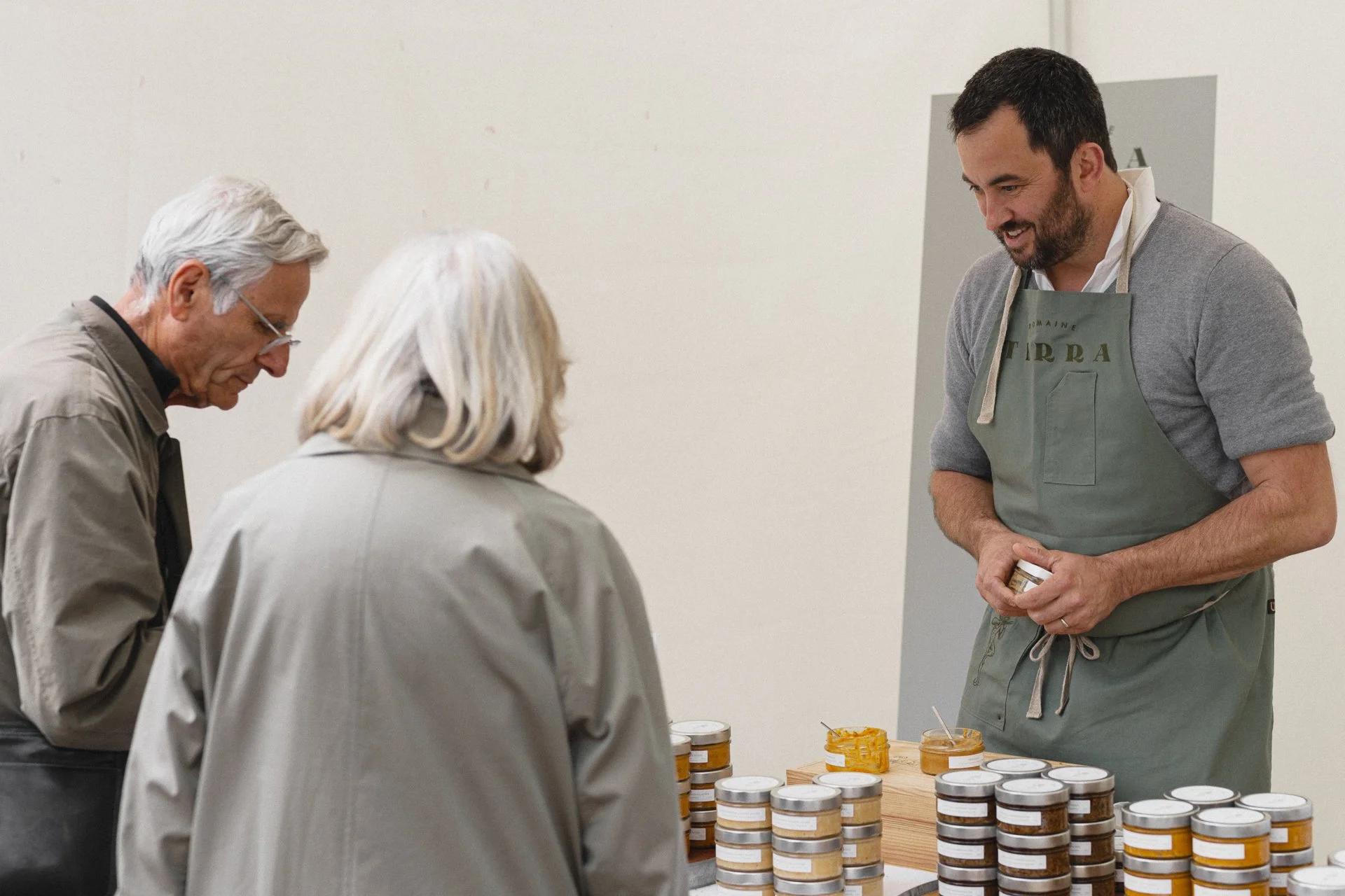 Un homme porte un tablier discutant avec deux personnes âgées devant des pots de confiture sur une table.