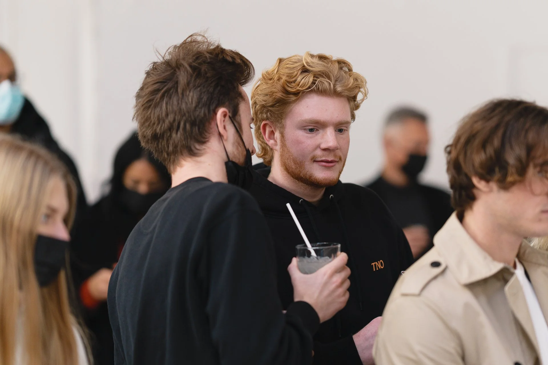 Jeune homme blond avec barbe portant un sweat noir, tenant un verre avec une paille, en conversation avec une personne portant un manteau beige et une chemise blanche, lors d'un rassemblement.