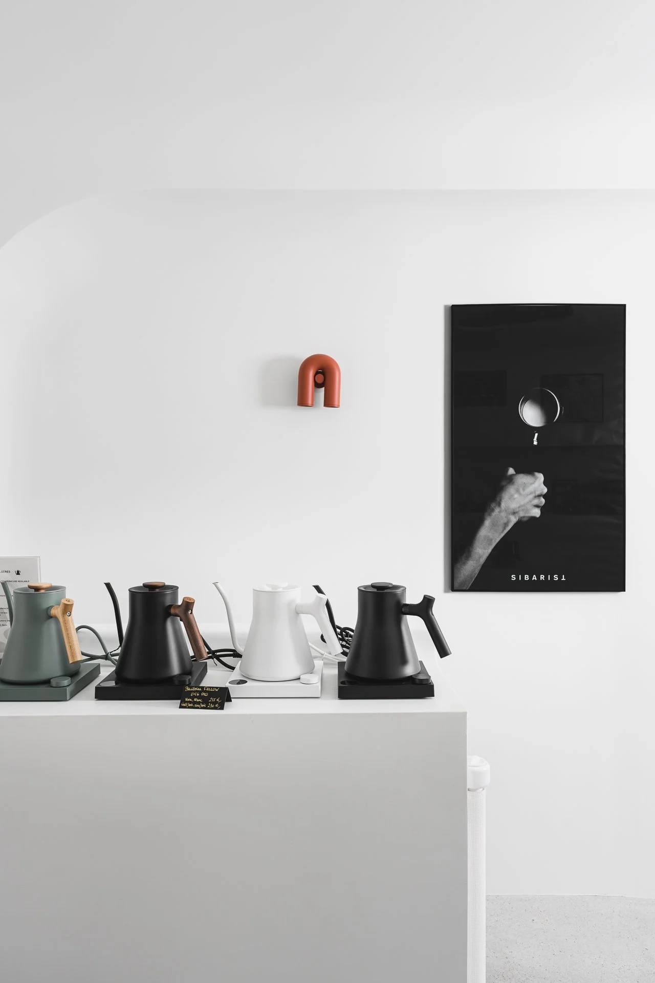 Une collection de cafetières colorées sur un comptoir blanc. Un tableau noir avec un doigt pointant vers le haut est accroché au mur blanc derrière, avec un affichage en français.