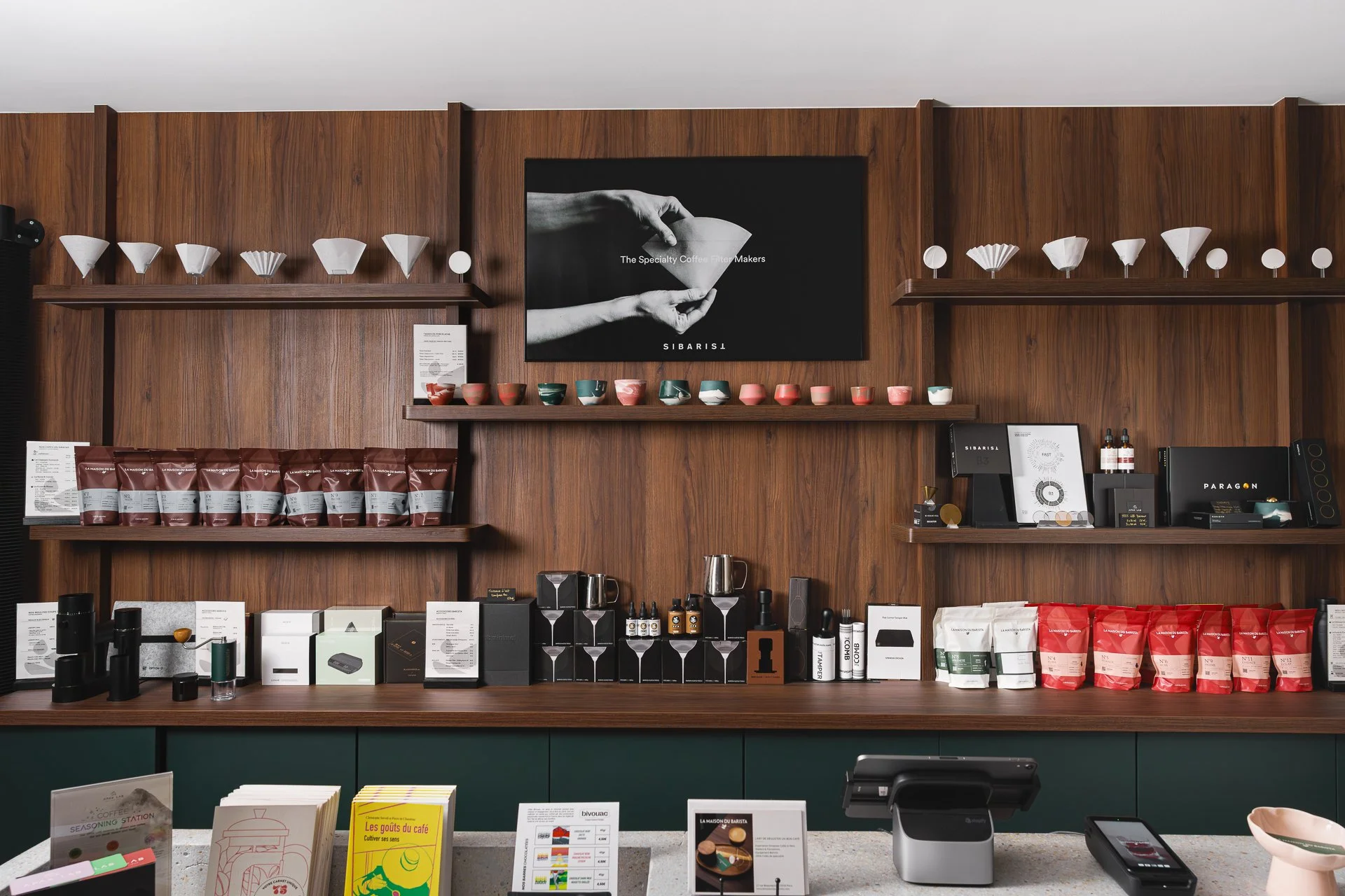 Espace intérieur d'un café ou boutique avec des étagères en bois affichant diverses tasses, paquets de café et accessoires. Un écran noir avec une image monochrome et du texte, comptoir avec caisses, menus et produits.