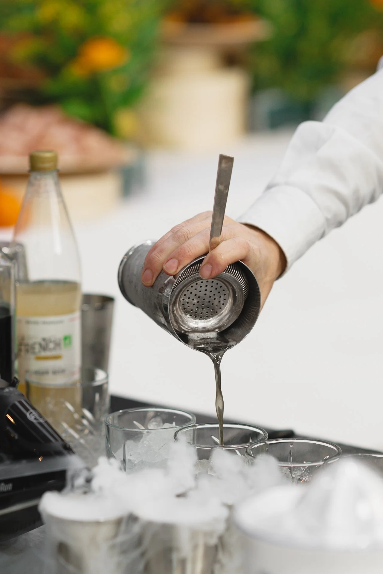 Une personne verse un liquide transparent à partir d'une bouteille en métal dans un verre, devant un fond flou avec des fruits et des plantes.