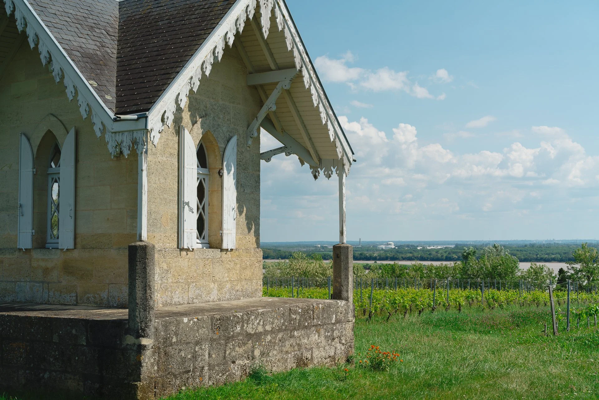 Une maison en pierre avec des volets en bois blanc et une toiture en ardoise, située dans un paysage rural avec un champ de vignes et un ciel en partie nuageux.