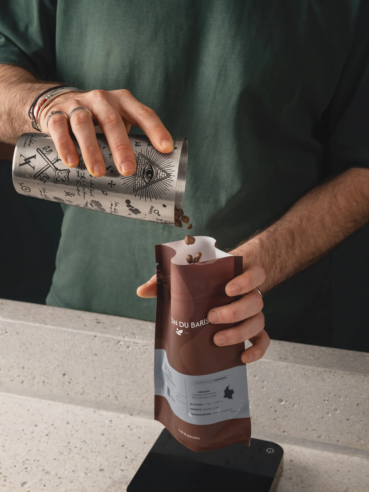 Une personne verse des grains de café moulus d'une tasse en métal décorée avec des symboles ésotériques dans un sac de café, portant un bracelet et des bagues, dans un environnement lumineux.