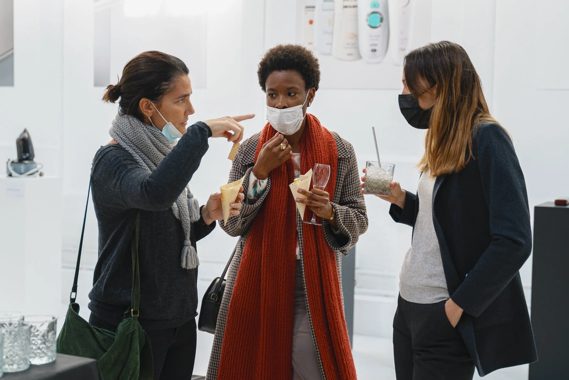 Trois femmes discutent dans une galerie d'art, portant des masques faciaux, en tenant des verres de boissons et une glace, pendant une exposition d'œuvres d'art.