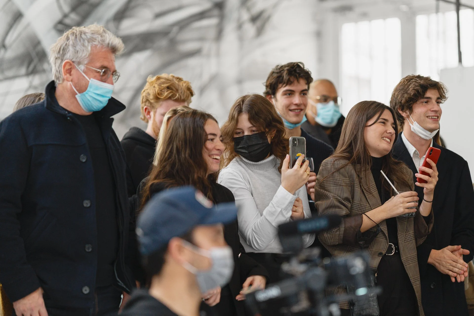 groupe de jeunes adultes, dont certains portent des masques faciaux, qui sourient et prennent des selfies dans un espace intérieur moderne avec des grandes fenêtres